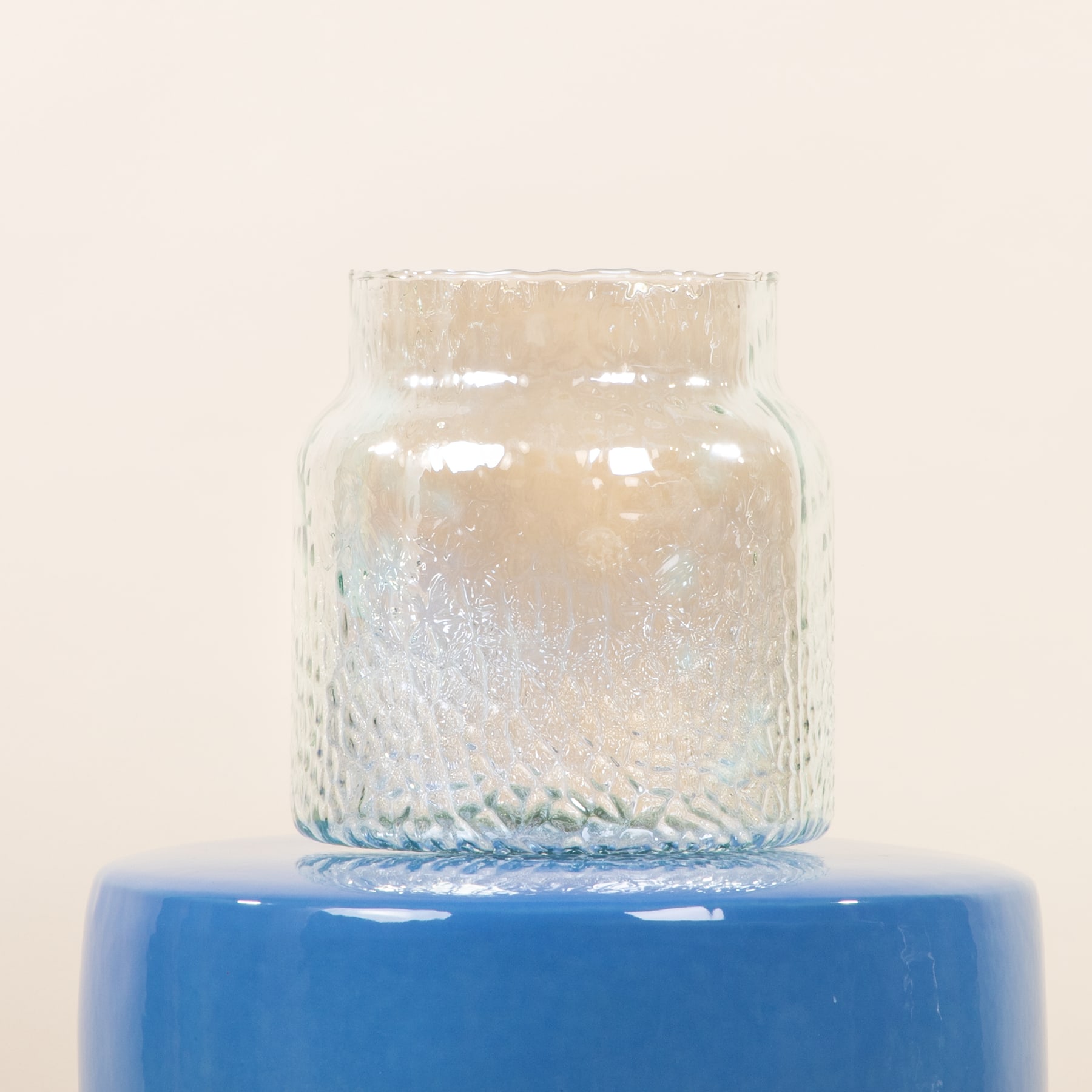 Eine Vase oder ein Gefäß aus klarem Glas mit strukturierter Oberfläche steht auf einer leuchtend blauen Plattform oder Oberfläche vor einem schlichten, hellen Hintergrund.