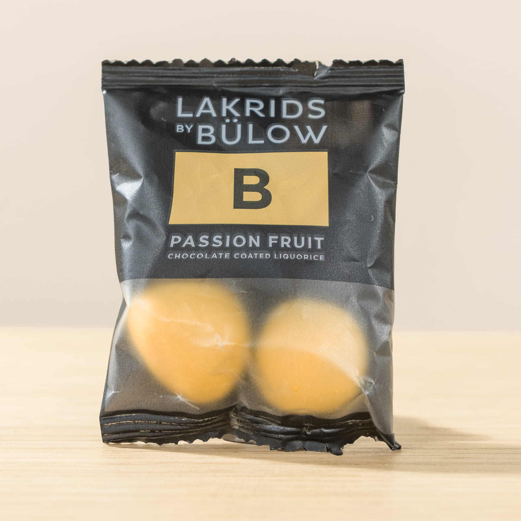 Eine Packung schokoladenüberzogener Lakritzbonbons von Lakrids by Bulow in der Geschmacksrichtung „Passionsfrucht“ liegt auf einer Holzoberfläche vor einem schlichten Hintergrund.