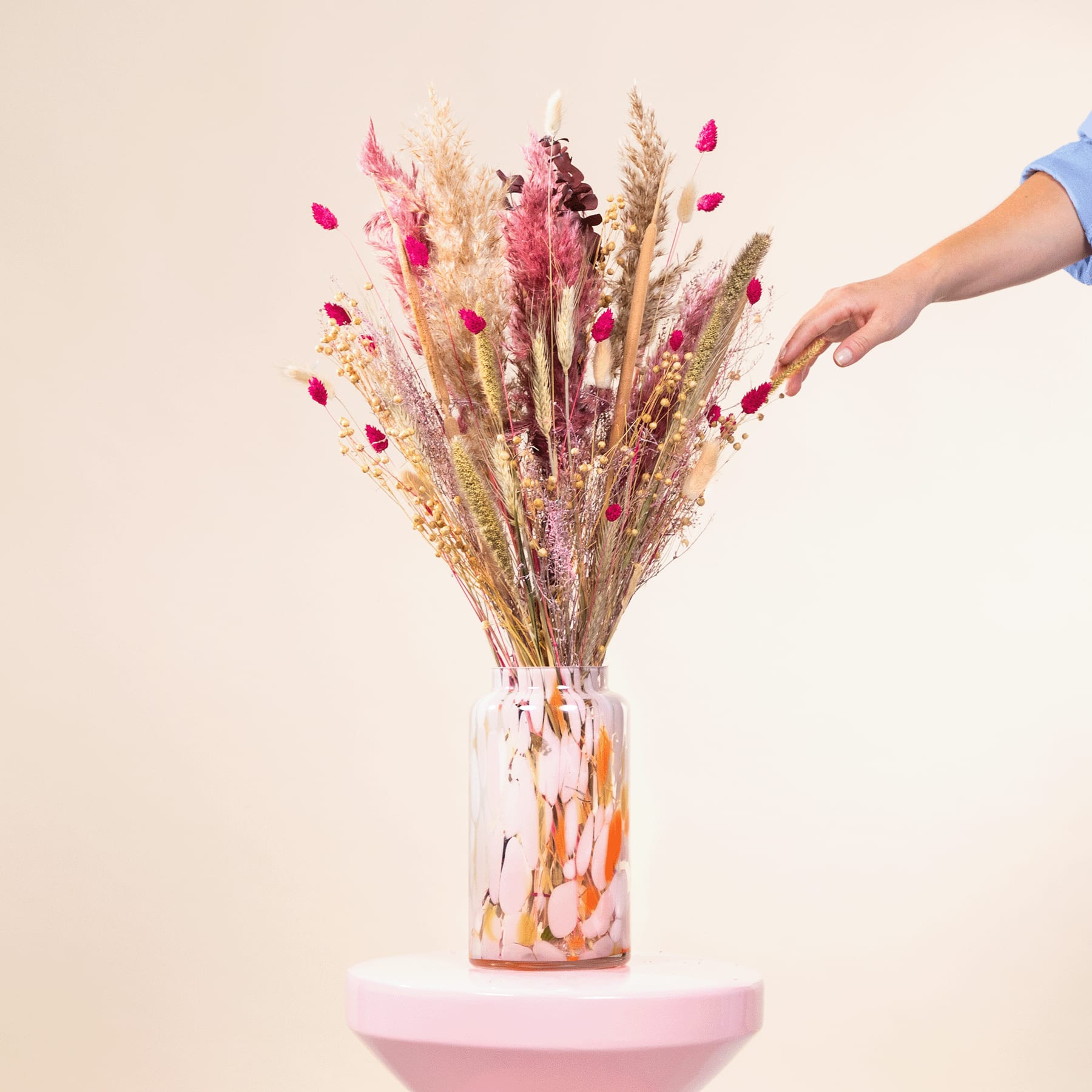 Ein farbenfroher Strauß getrockneter Blumen und Blütenblätter in einer Glasvase vor einem schlichten beigen Hintergrund, in den eine Hand hineingreift, um das Arrangement zu richten.