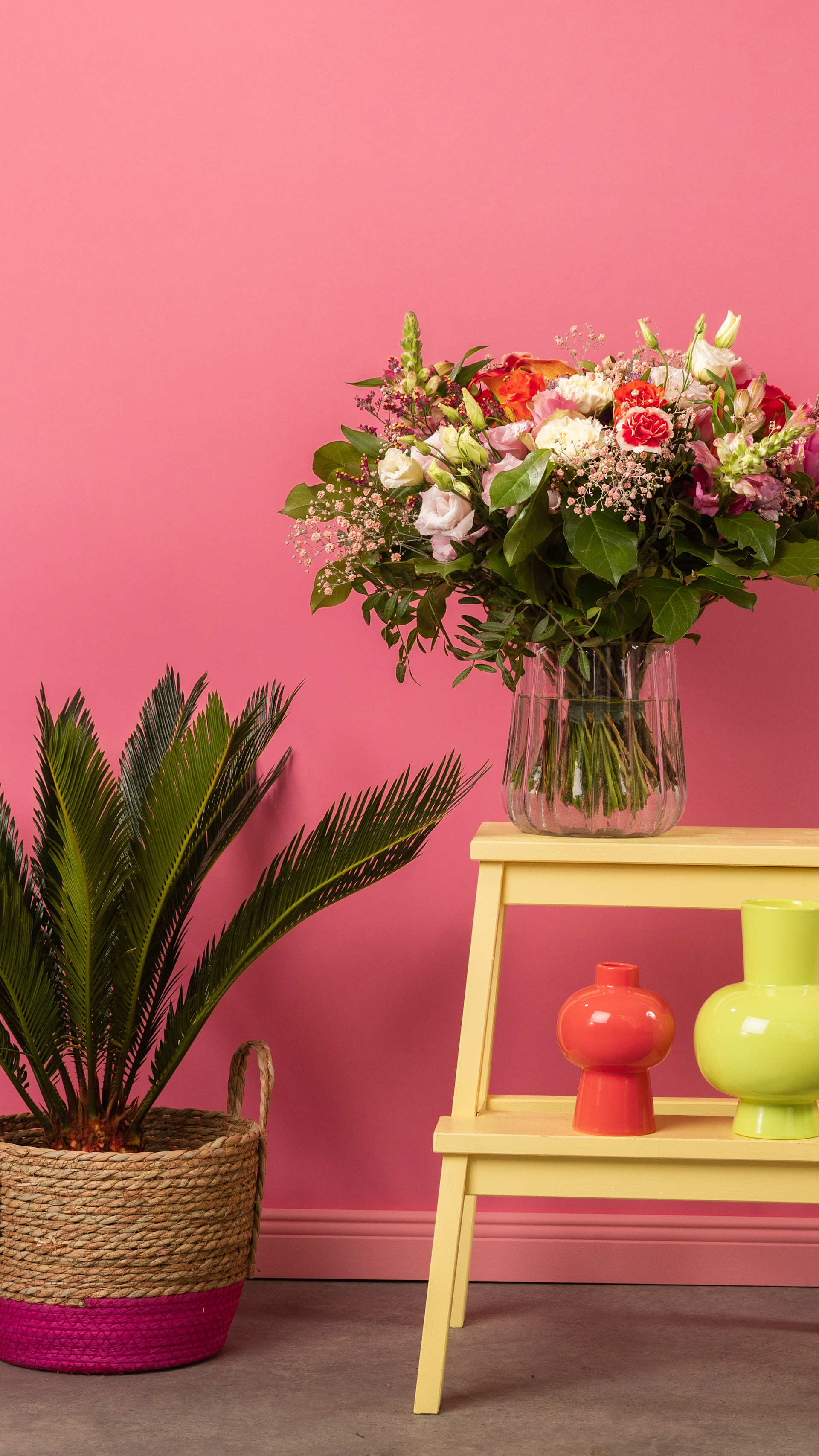 Auf einem gelben Holztisch steht ein farbenfrohes Blumenarrangement in einer Glasvase, begleitet von einer Topfpflanze und Dekorationsobjekten vor einem leuchtend rosa Hintergrund.