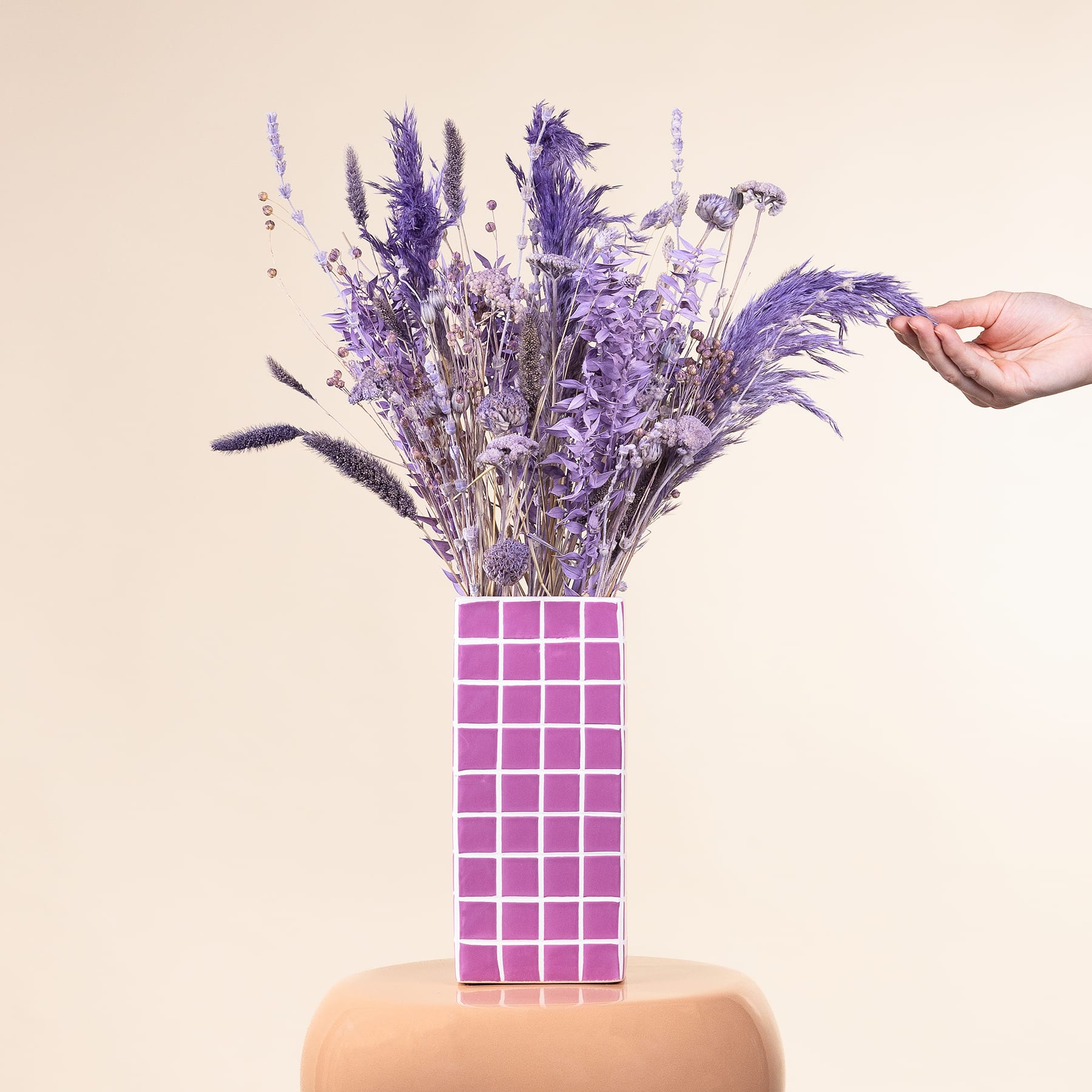 Eine Hand hält einen leuchtenden Strauß lila Blumen in einer Vase mit einem rosa-weißen Schachbrettmuster vor einem neutralen Hintergrund.