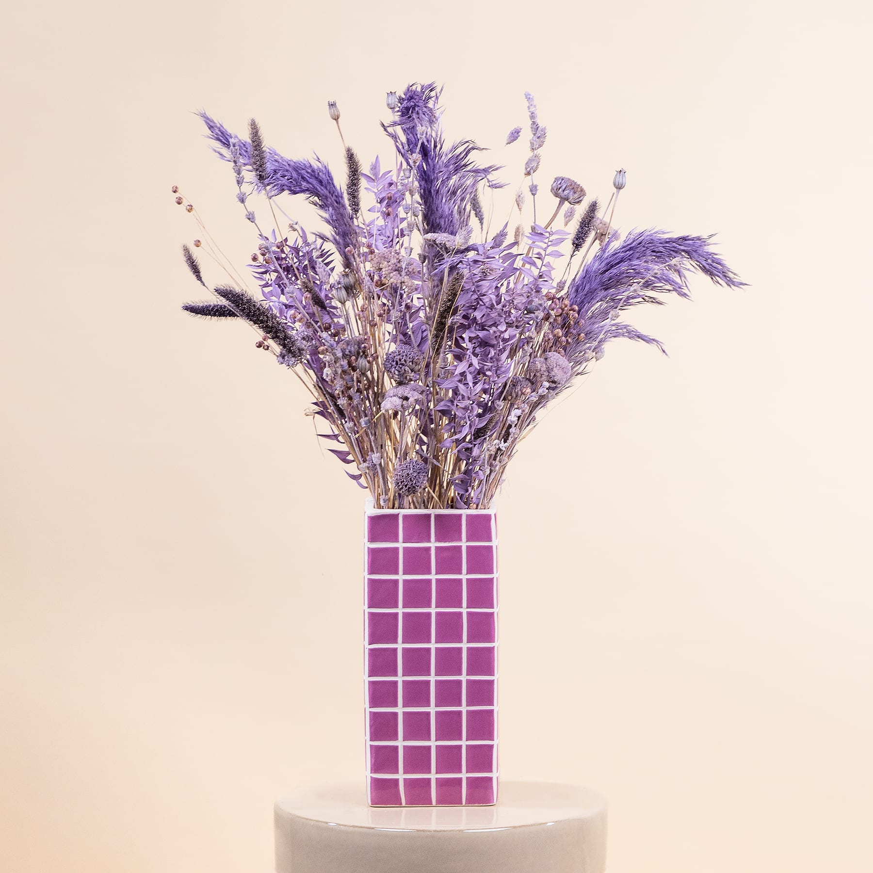 Ein farbenfroher Strauß violetter Blumen, darunter Lavendel und andere zarte Blüten, wird in einer geometrischen rosa Vase vor einem neutralen beigen Hintergrund präsentiert.