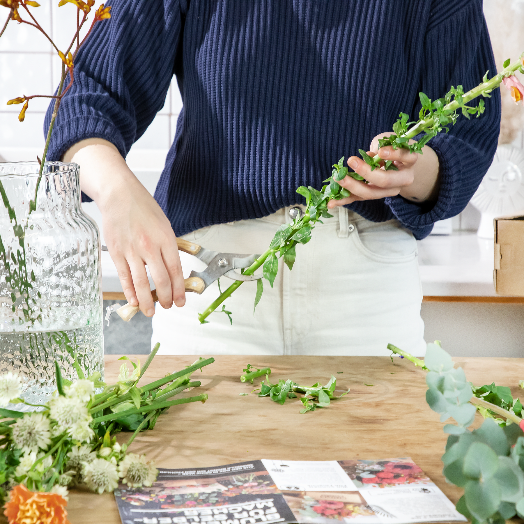 Eine Person in einem blau gestreiften Hemd arrangiert frische grüne Blätter und Blumen auf einer Holzoberfläche, im Hintergrund sind verschiedene andere florale Elemente zu sehen.