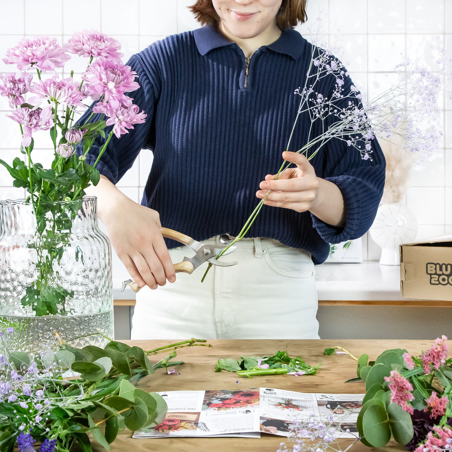 Eine Frau in einem marineblauen Pullover arrangiert einen Strauß rosa Blumen auf einem Holztisch, umgeben von weiteren Blumenarrangements und Büchern.