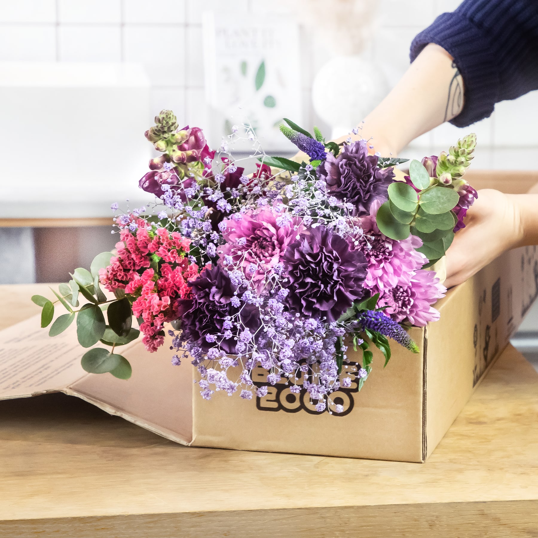 Ein farbenfroher Strauß violetter, rosa und weißer Blumen ist in einer Holzkiste arrangiert und steht auf einer Holzoberfläche, im Hintergrund sind ein Fenster und helle Wände zu sehen.