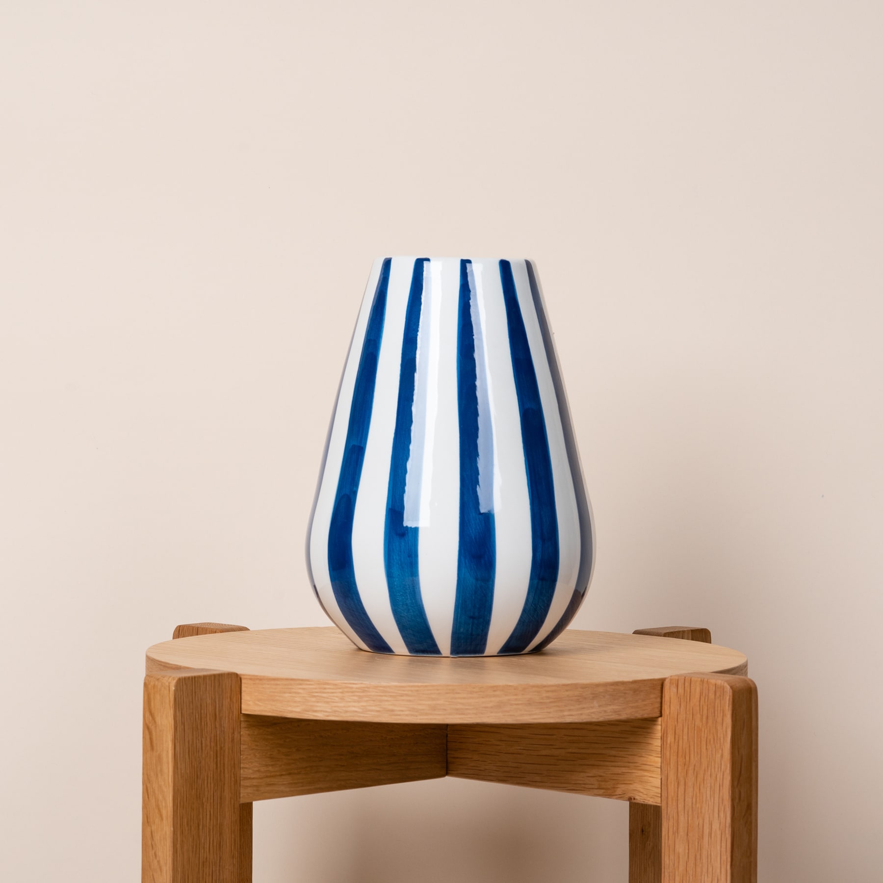 Eine blau-weiß gestreifte Keramikvase steht auf einem Holzständer vor einem schlichten beigen Hintergrund.