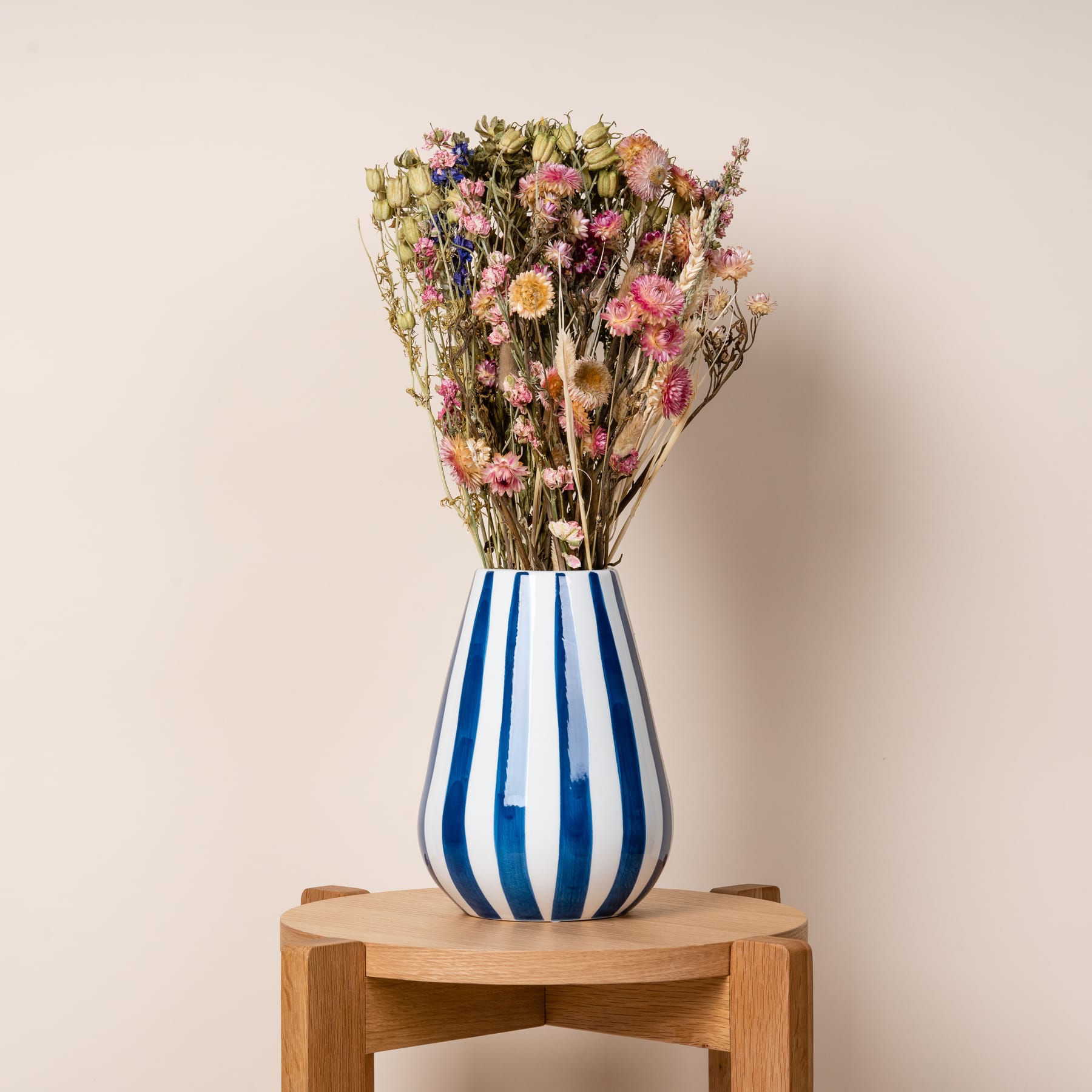 Ein Strauß getrockneter Blumen in einer blau-weiß gestreiften Keramikvase steht auf einem Holzständer vor einem beigen Hintergrund.