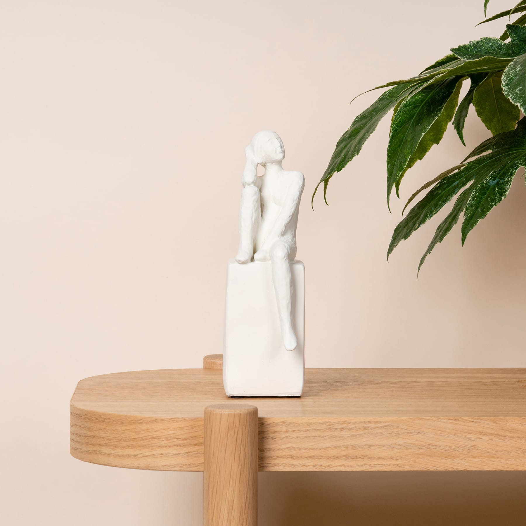 Eine kleine weiße Keramikfigur steht auf einer Holzplattform, im Hintergrund eine üppige grüne Pflanze.
