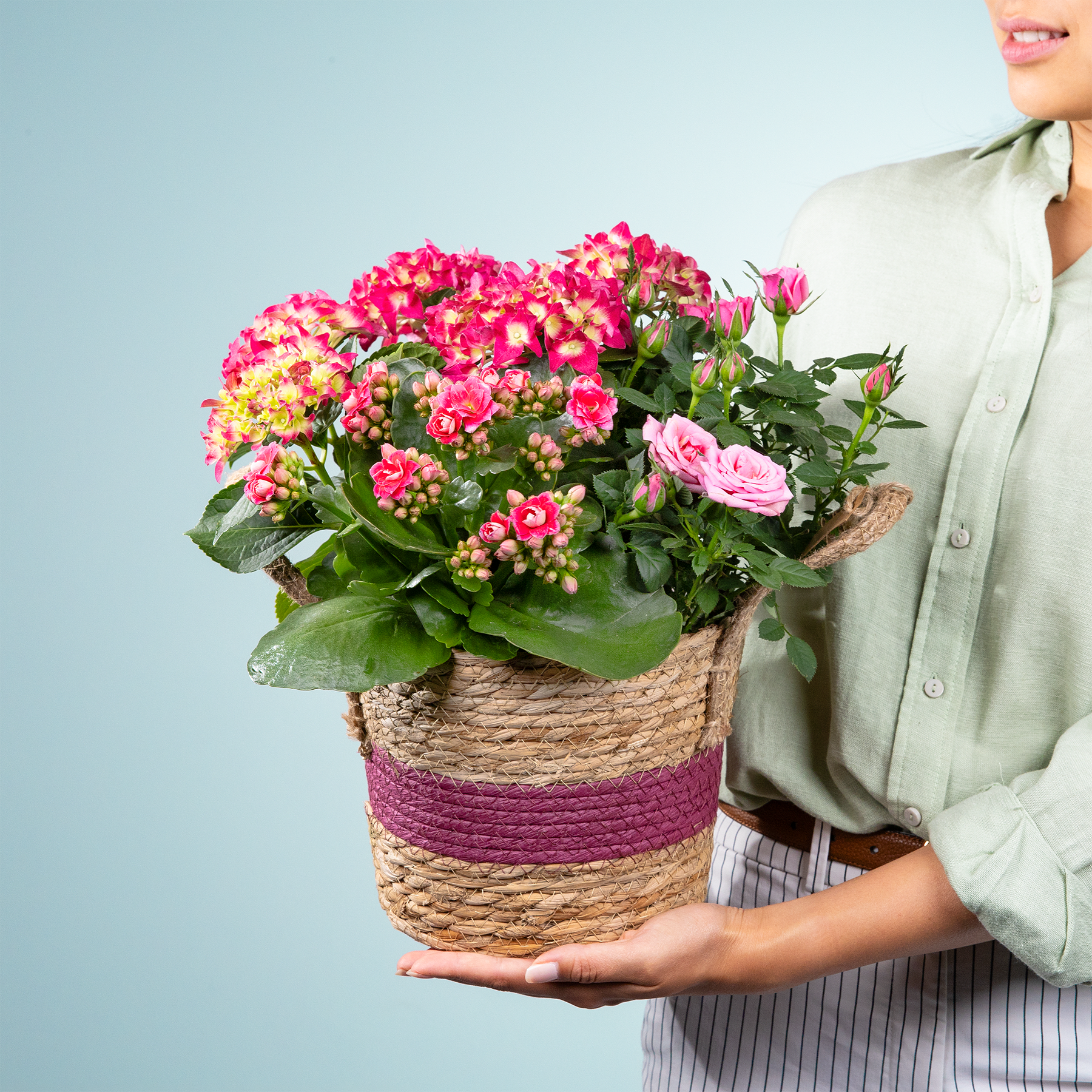 Eine Person hält einen farbenfrohen Strauß rosa und gelber Blumen in einem geflochtenen Korb vor einem schlichten blauen Hintergrund.