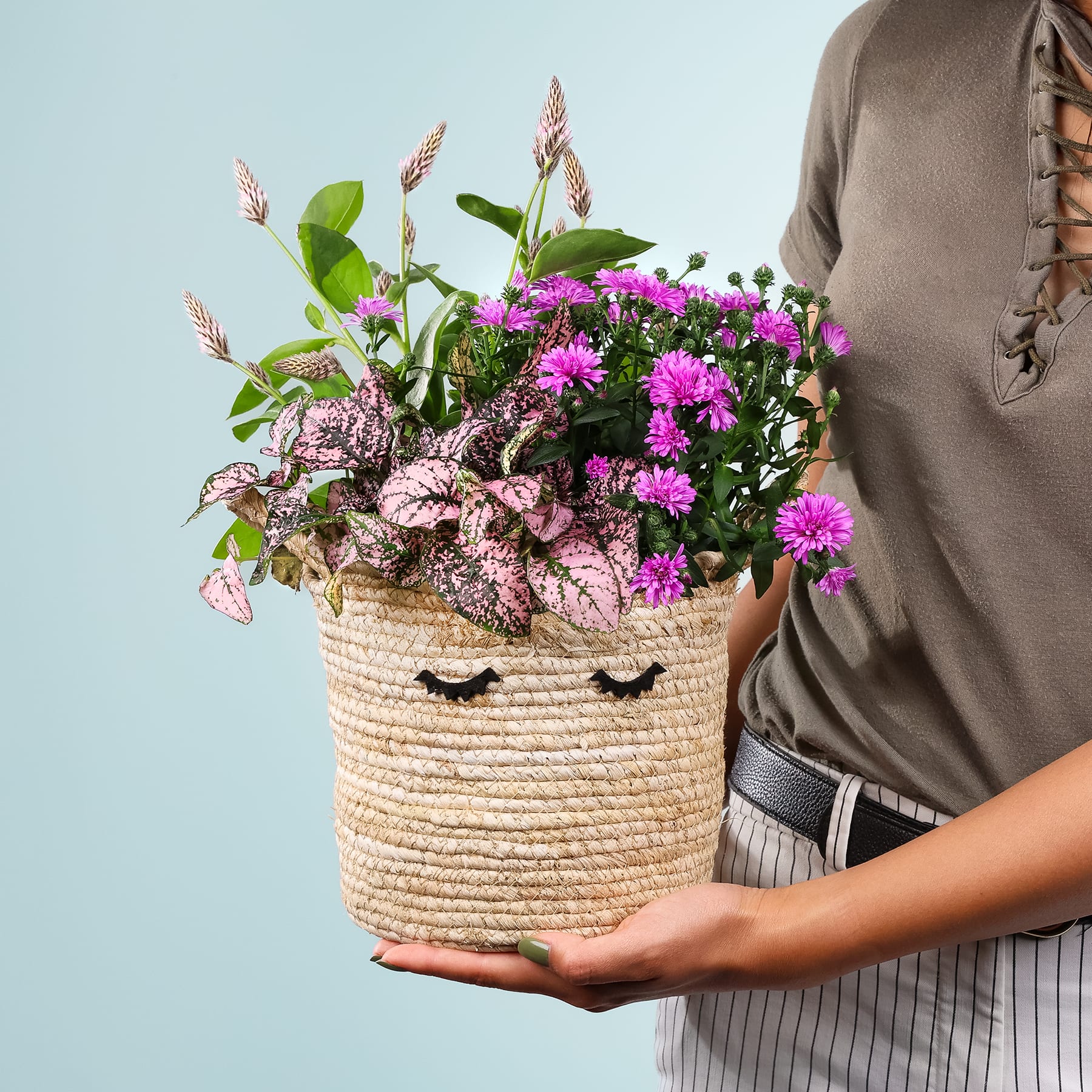 Eine Person hält einen geflochtenen Korb voller leuchtend rosa und violetter Blumen vor einem hellblauen Hintergrund.