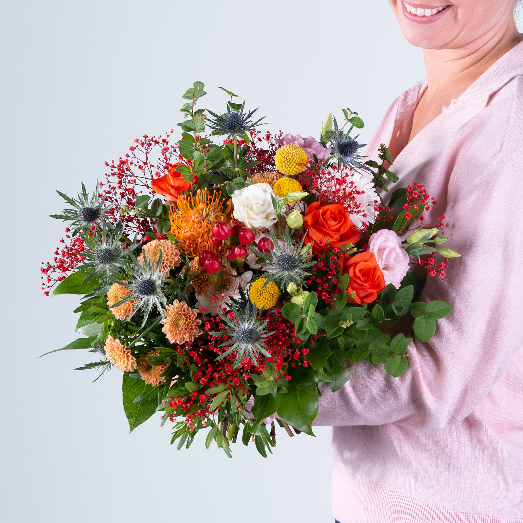 Ein leuchtender Strauß bunter Blumen, darunter Rosen, Chrysanthemen und andere Blüten, wird von einer Person gehalten, die einen rosa Pullover vor einem schlichten Hintergrund trägt.