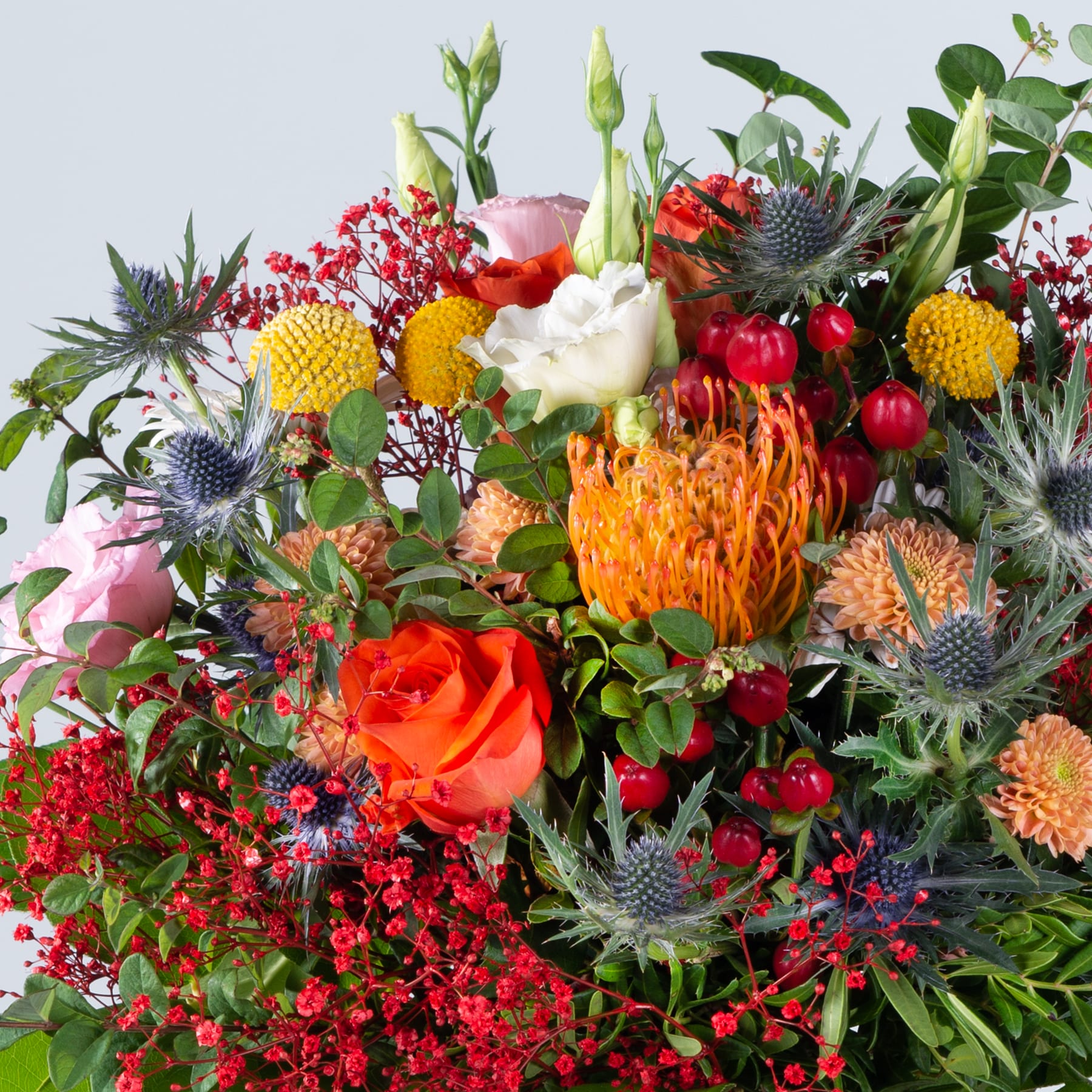 Ein lebendiger und üppiger Strauß verschiedener bunter Blumen, darunter Rosen, Proteas und Disteln, arrangiert in einer rustikalen, natürlich wirkenden Anordnung vor einem schlichten Hintergrund.