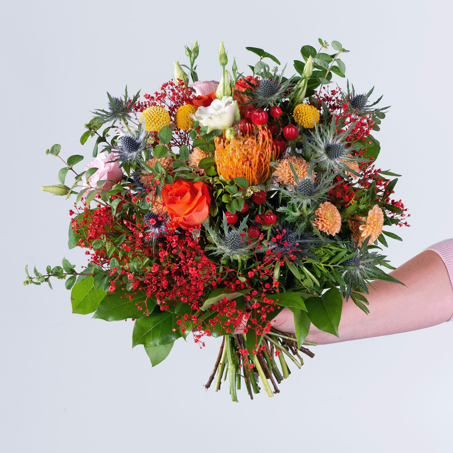 Ein lebendiger und üppiger Strauß verschiedener bunter Blumen, darunter Rosen, Proteen und andere exotische Blüten, die eine Person vor einem schlichten Hintergrund in den Händen hält.