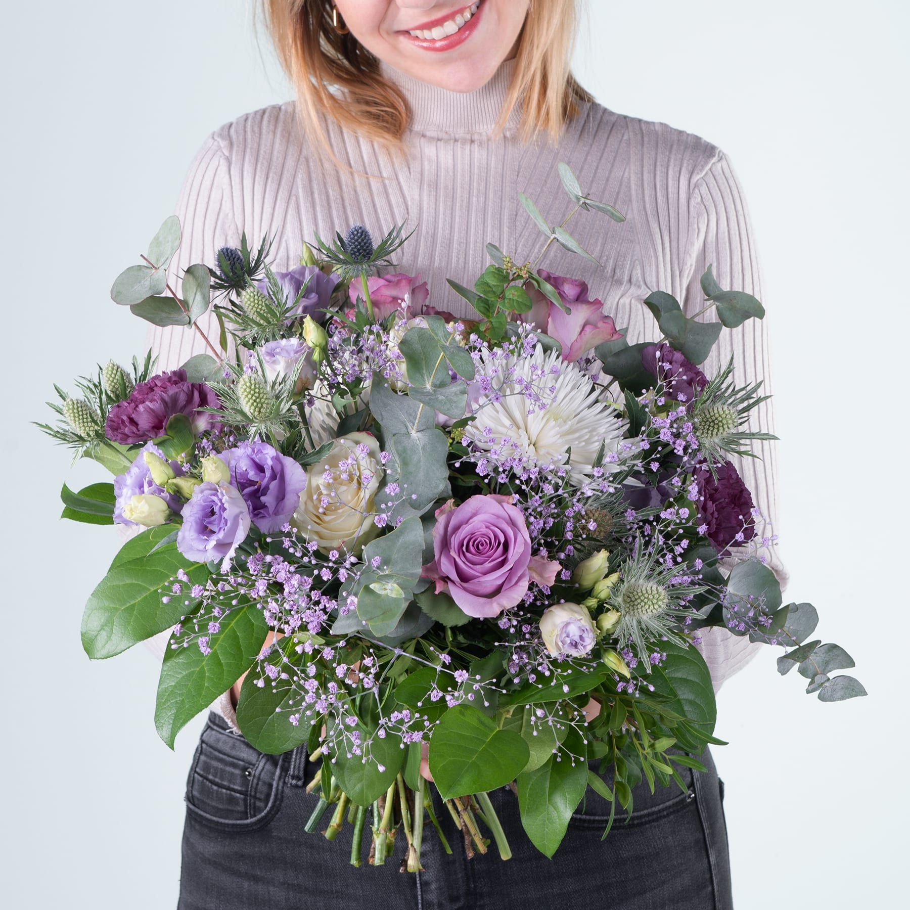 Eine lächelnde Frau hält einen farbenfrohen Strauß lila, rosa und weißer Blumen vor einem schlichten Hintergrund.