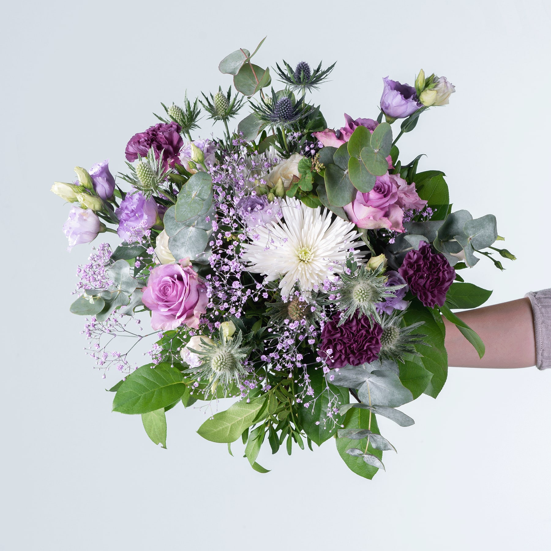 Ein farbenfroher Strauß verschiedener Blumen, darunter violette und rosa Rosen, weiße Gänseblümchen und Grünpflanzen, arrangiert in einer Vase vor einem schlichten, hellen Hintergrund.