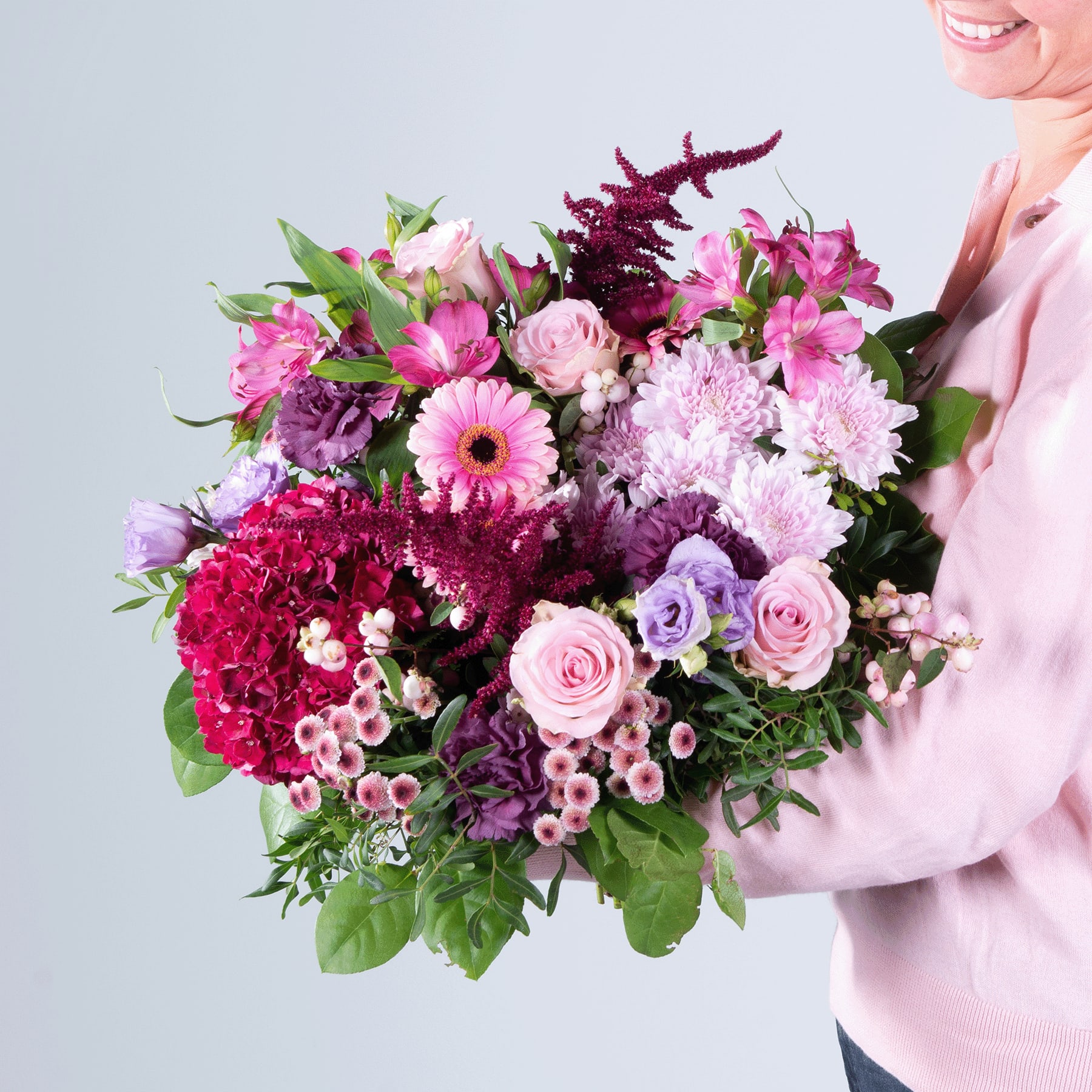 Ein farbenfroher Strauß verschiedener rosa, violetter und roter Blumen, darunter Rosen, Gerbera und Chrysanthemen, wird von einer Person in einem rosa Pullover vor einem schlichten Hintergrund gehalten.