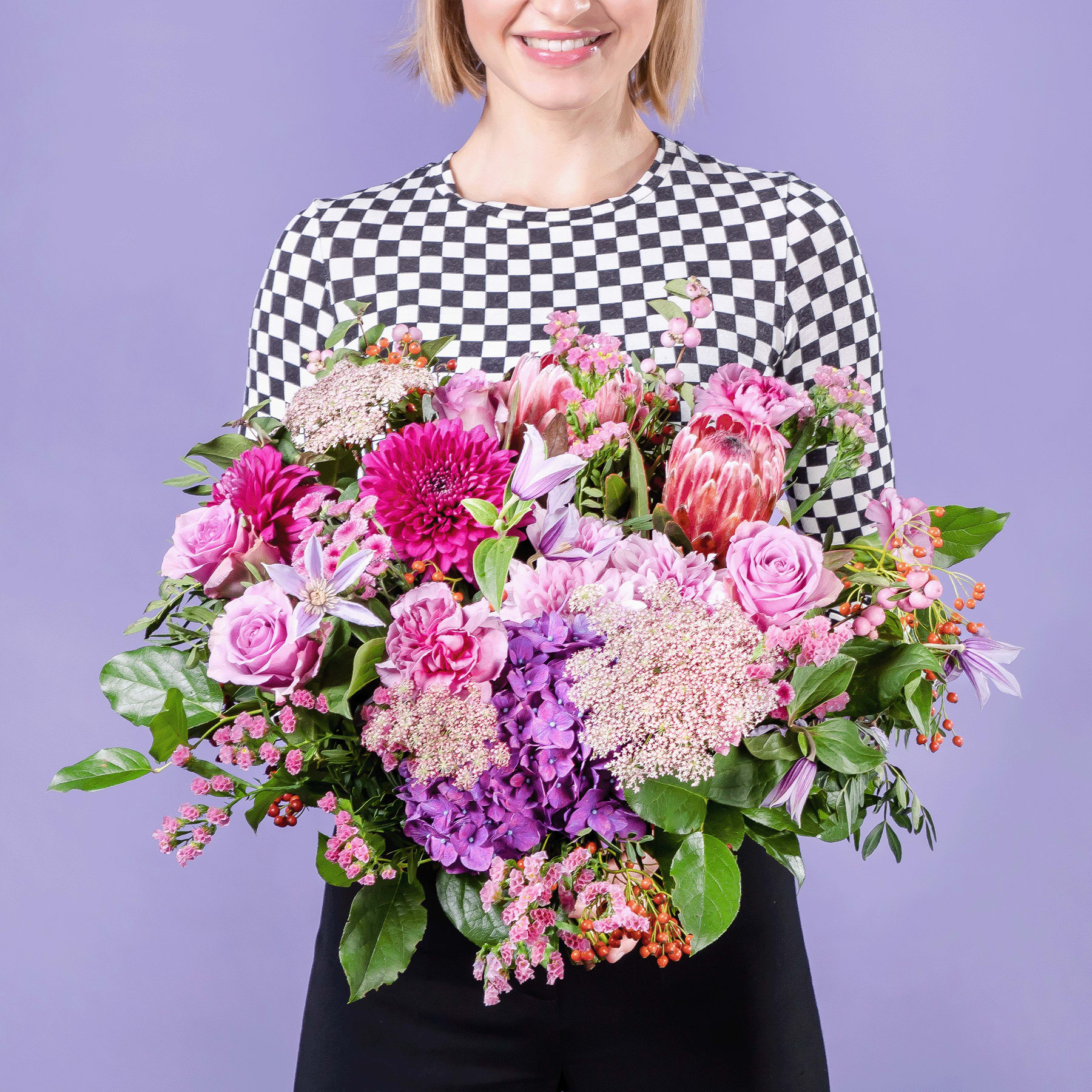 Eine lächelnde Frau in einem karierten Oberteil hält einen farbenfrohen Strauß rosa, violetter und weißer Blumen vor einem einfarbig violetten Hintergrund.