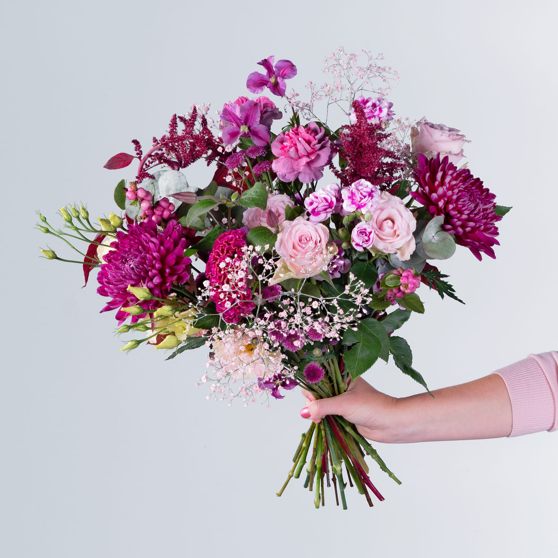 Ein farbenfroher Strauß verschiedener rosa und violetter Blumen, darunter Rosen, Dahlien und Orchideen, wird vor einem schlichten Hintergrund in den Händen einer Person gehalten.