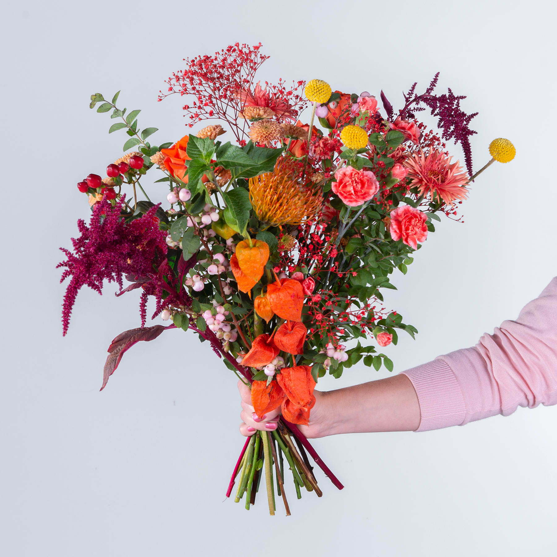 Ein leuchtender Strauß verschiedener bunter Blumen, darunter Rosen, Chrysanthemen und andere Blüten, den eine Person vor einem schlichten Hintergrund in den Händen hält.