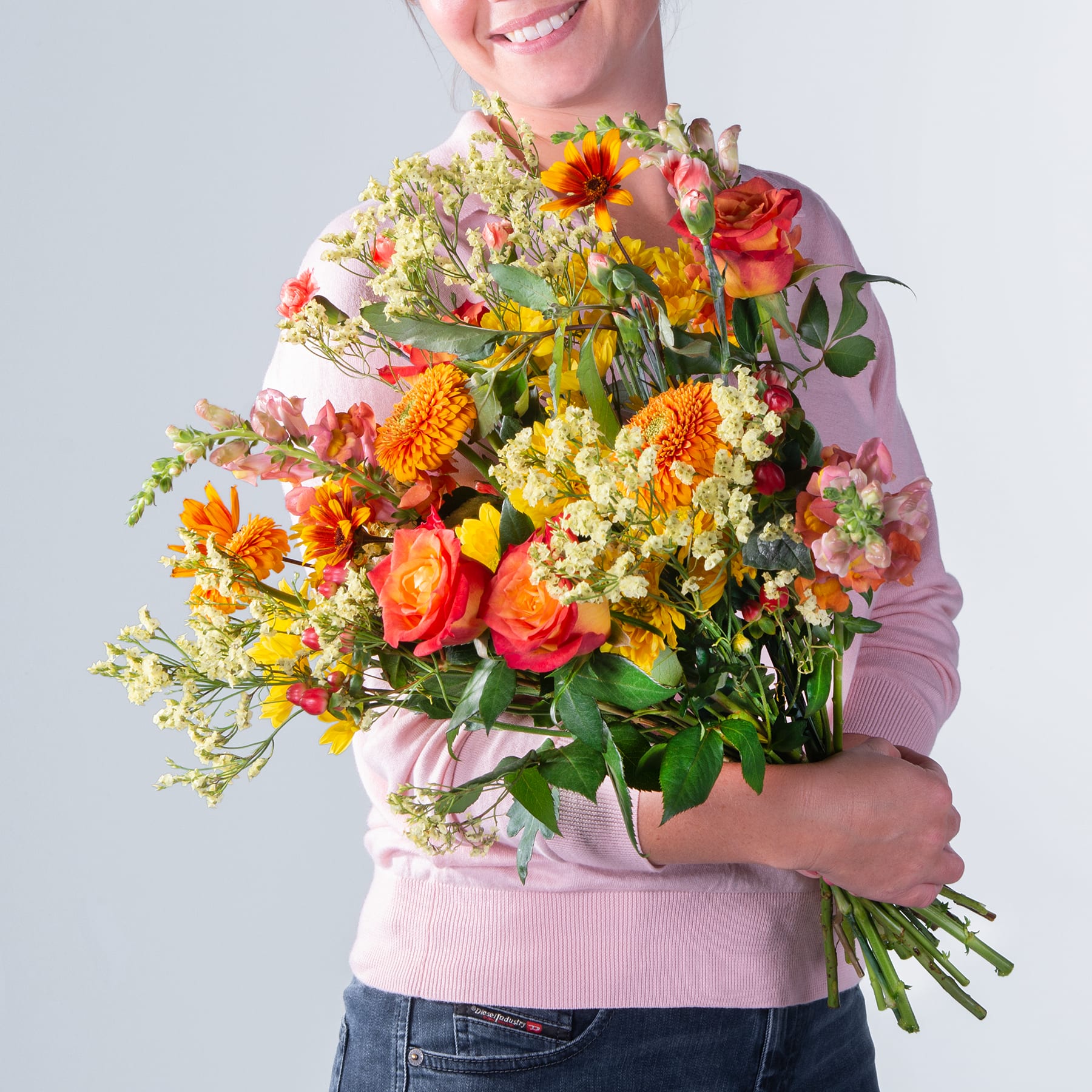 Eine lächelnde Frau in einem rosa Pullover hält einen großen Strauß leuchtend bunter Blumen vor einem schlichten grauen Hintergrund.