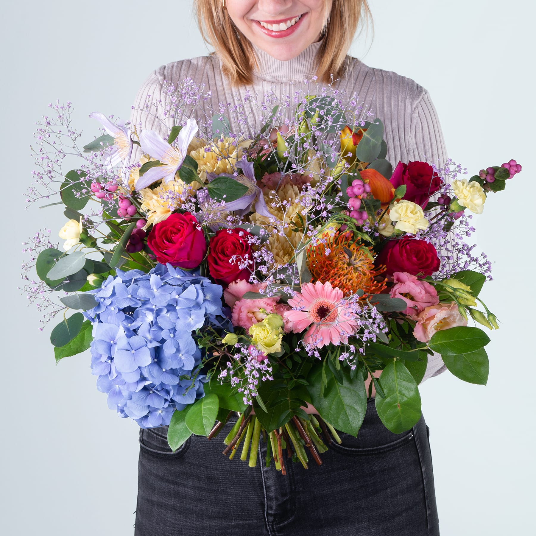 Eine lächelnde Frau hält vor einem schlichten Hintergrund einen leuchtend bunten Blumenstrauß, darunter rote Rosen, blaue Hortensien und verschiedene andere Blüten.