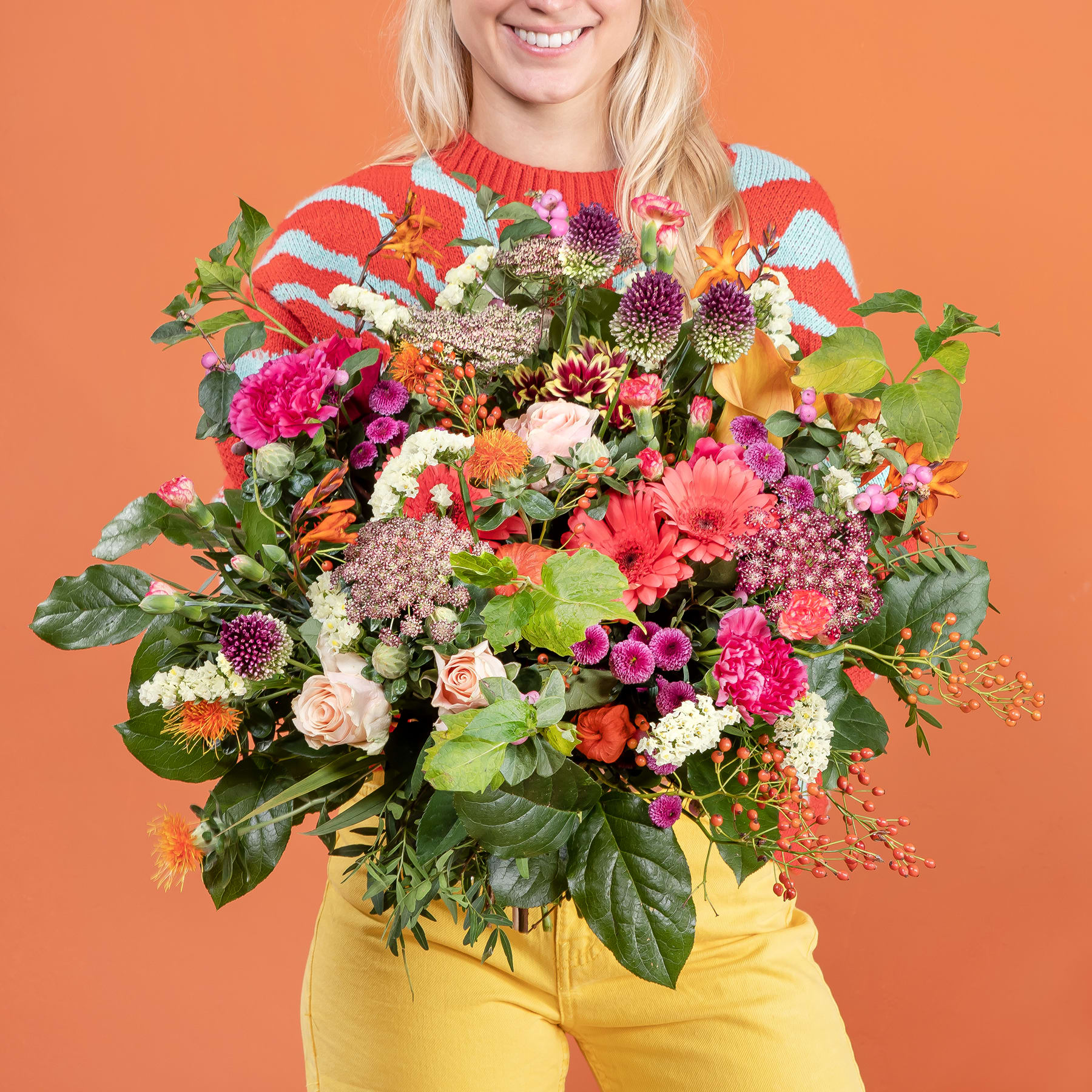 Ein leuchtender Strauß bunter Blumen, darunter Rosen, Dahlien und Grünpflanzen, wird von einer lächelnden Frau in einem gestreiften Pullover vor einem orangefarbenen Hintergrund gehalten.