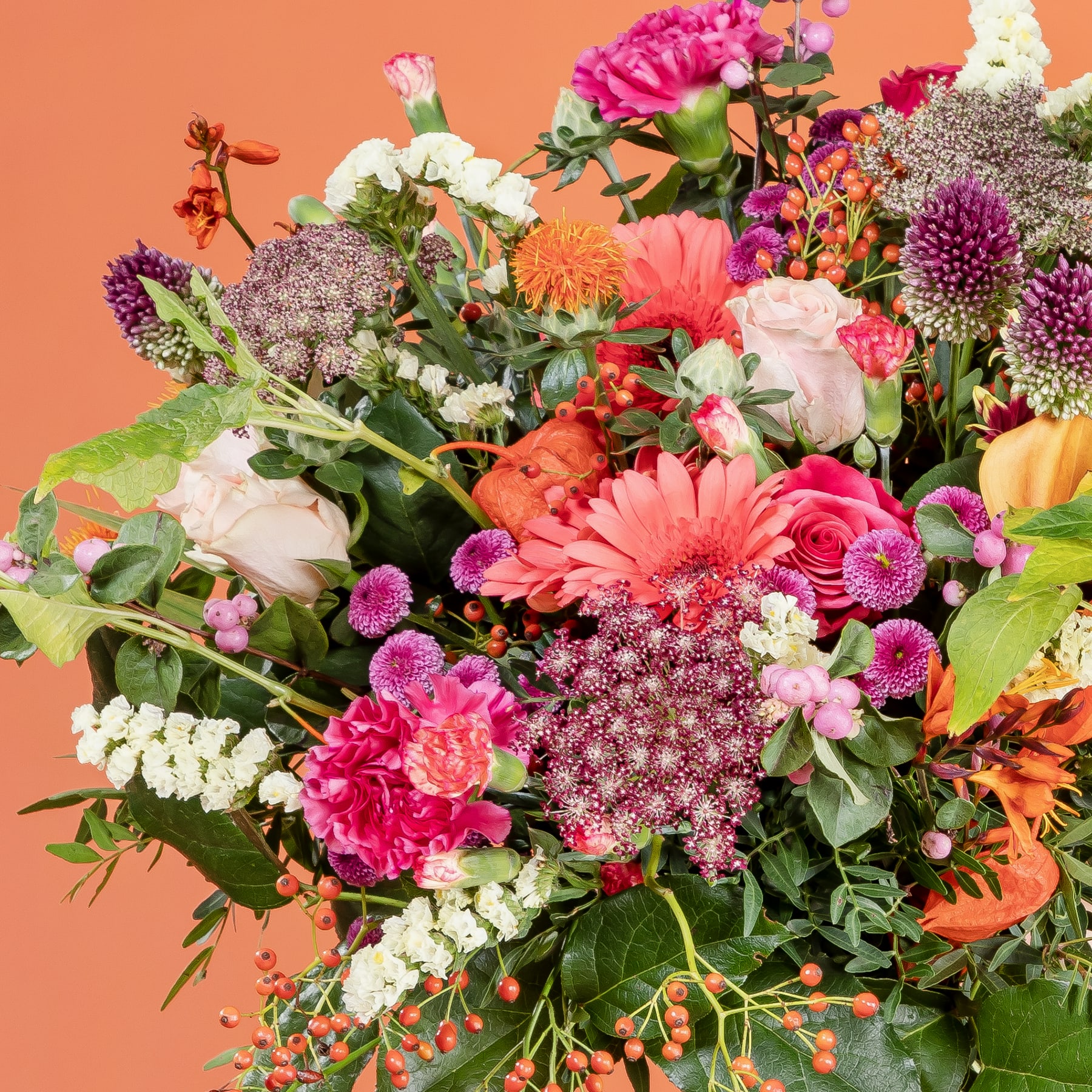 Ein lebendiger und üppiger Strauß verschiedener bunter Blumen, darunter rosa Gerbera, rote Nelken und violette Chrysanthemen, vor einem warmen orangefarbenen Hintergrund.