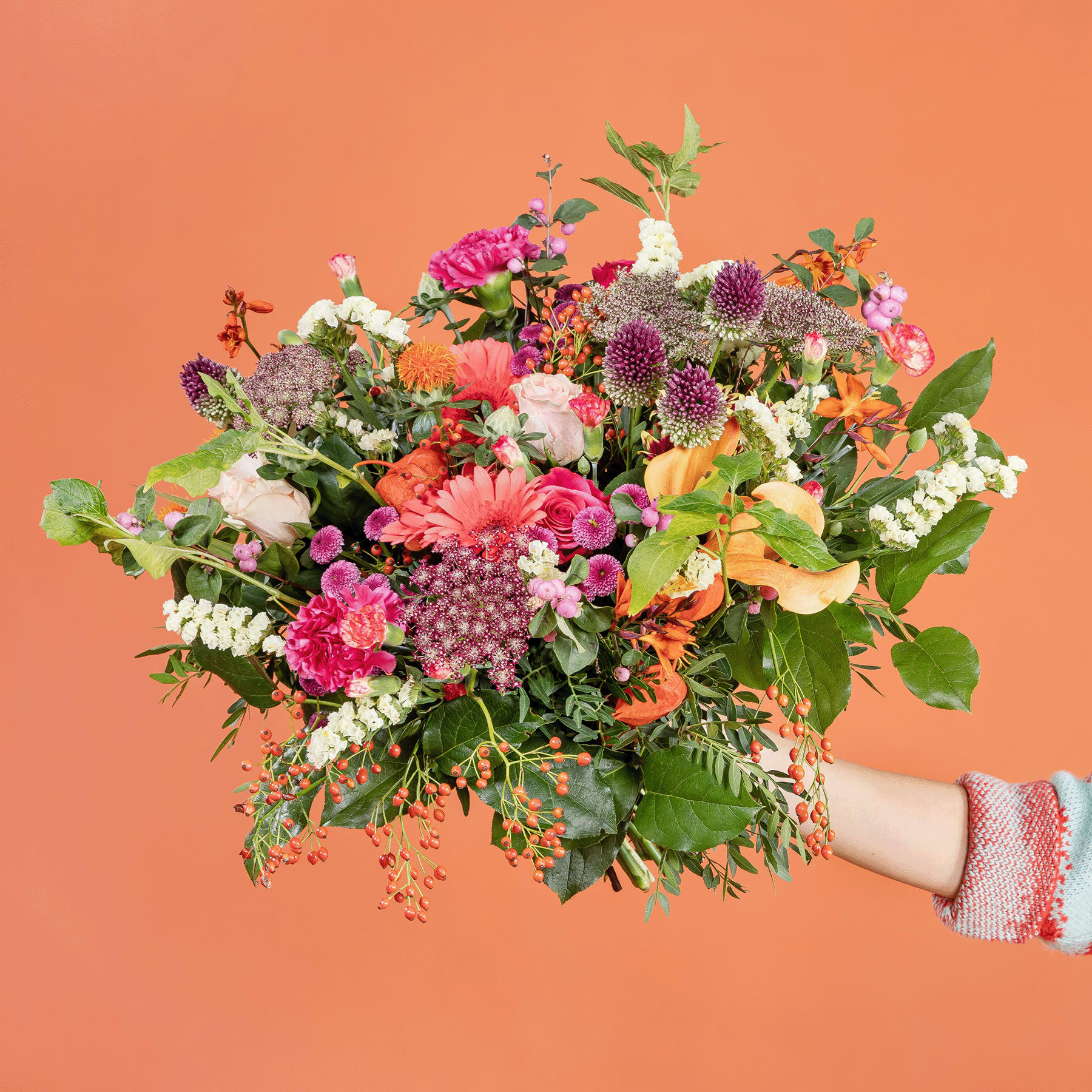 Ein leuchtender Strauß bunter Blumen, darunter rosa Gerbera, lila Allium und verschiedene andere Blüten, wird vor einem orangefarbenen Hintergrund in den Händen einer Person gehalten.
