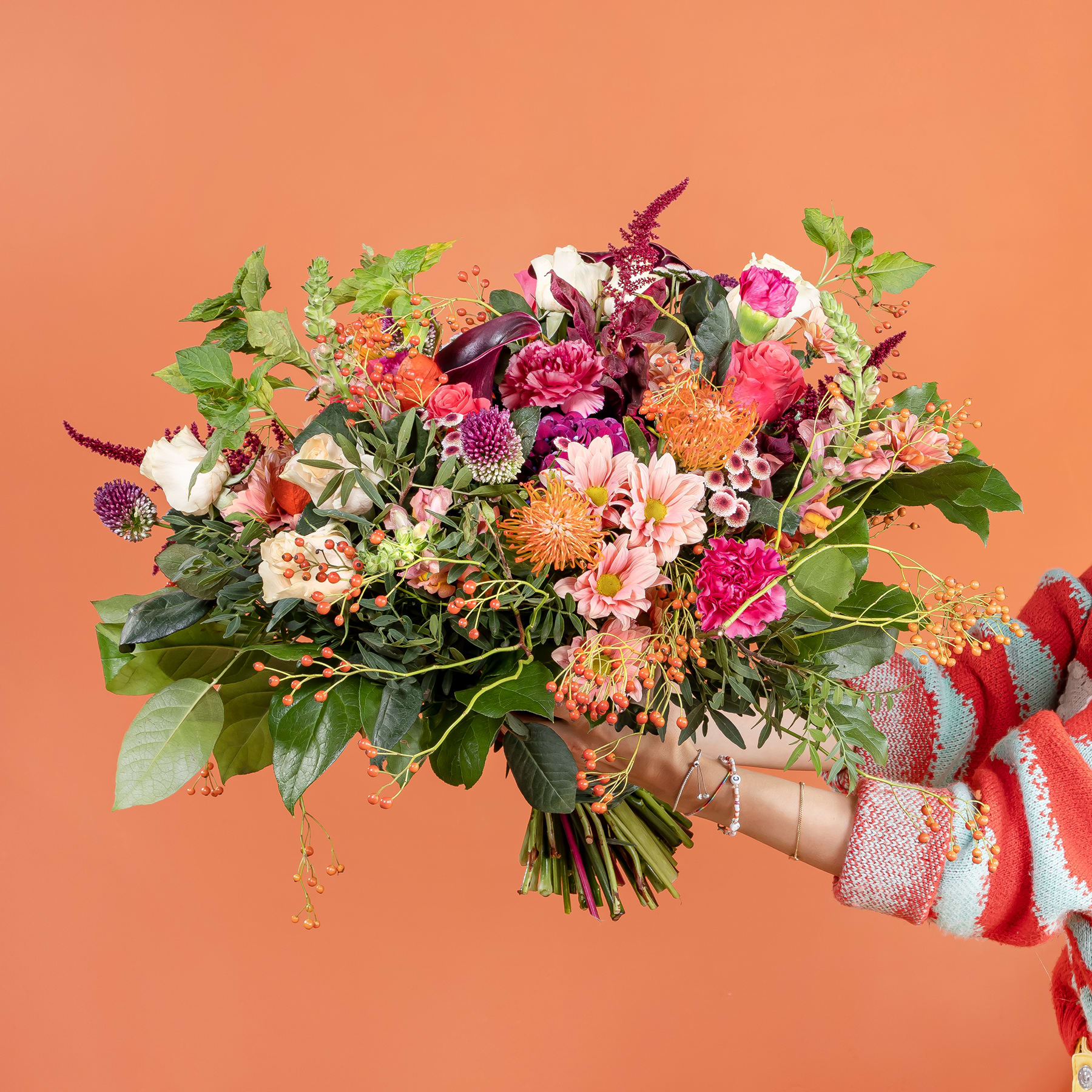 Ein lebendiger Strauß bunter Blumen, darunter Rosen, Dahlien und andere Blüten, gehalten in einer in einen gemusterten Schal gehüllten Hand vor einem warmen orangefarbenen Hintergrund.