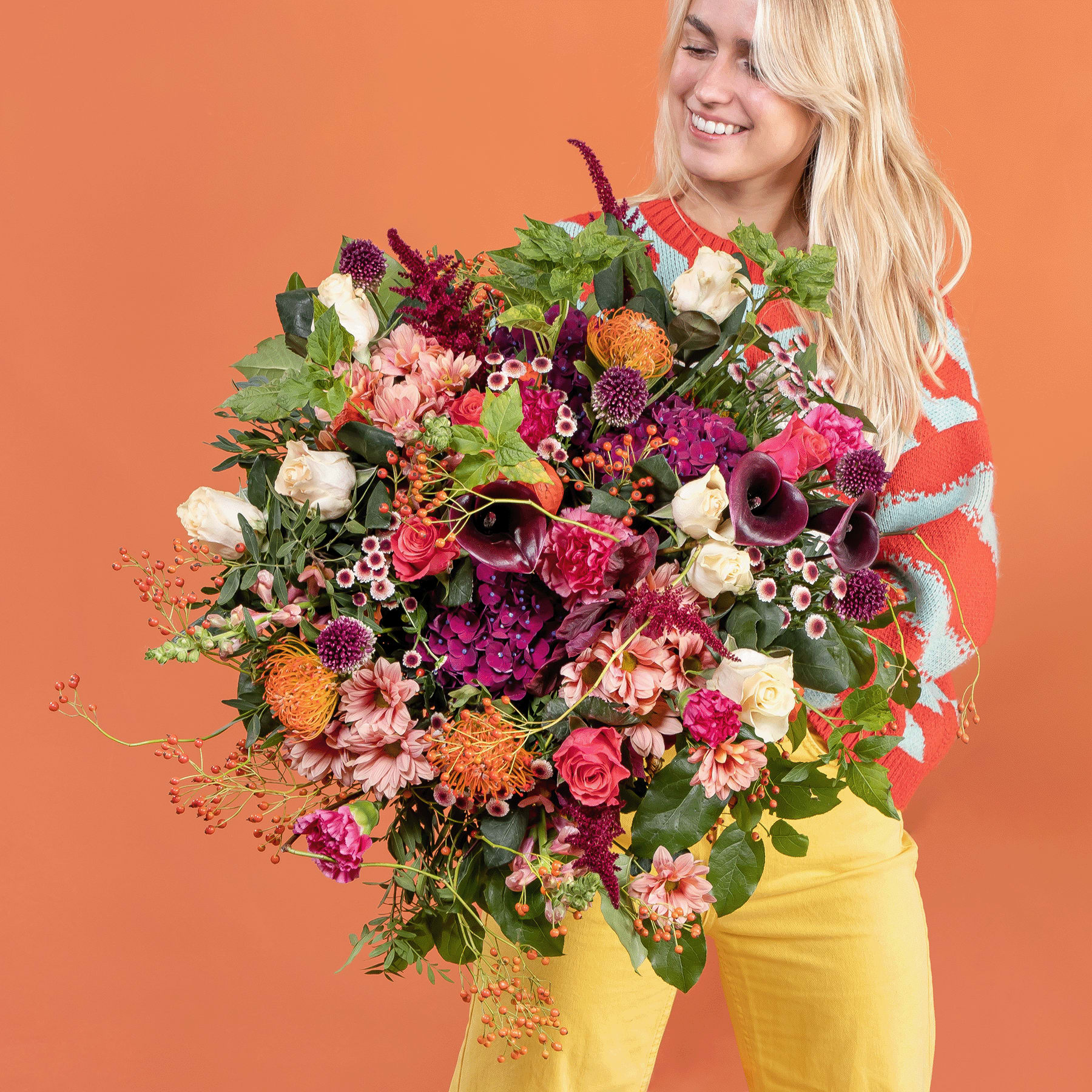 Ein leuchtender Strauß bunter Blumen, darunter Rosen, Gänseblümchen und andere Blüten, wird von einer lächelnden Frau gehalten, die ein rotes Oberteil und eine gelbe Hose vor einem orangefarbenen Hintergrund trägt.