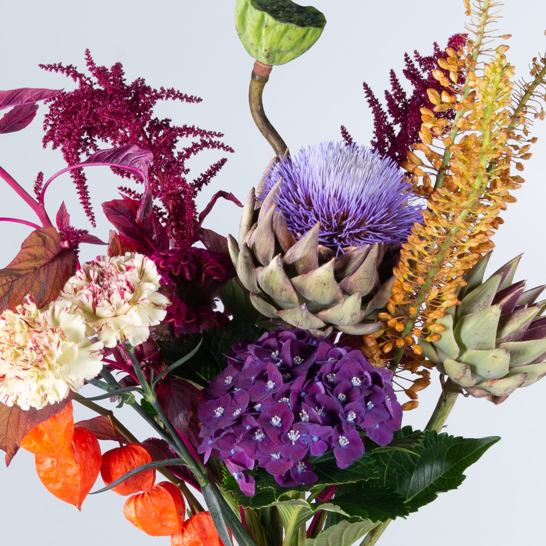 Ein lebendiger Strauß verschiedener bunter Blumen, darunter violette Artischocken, roter Amarant, gelbe Chrysanthemen und grünes Laub, vor einem schlichten Hintergrund.