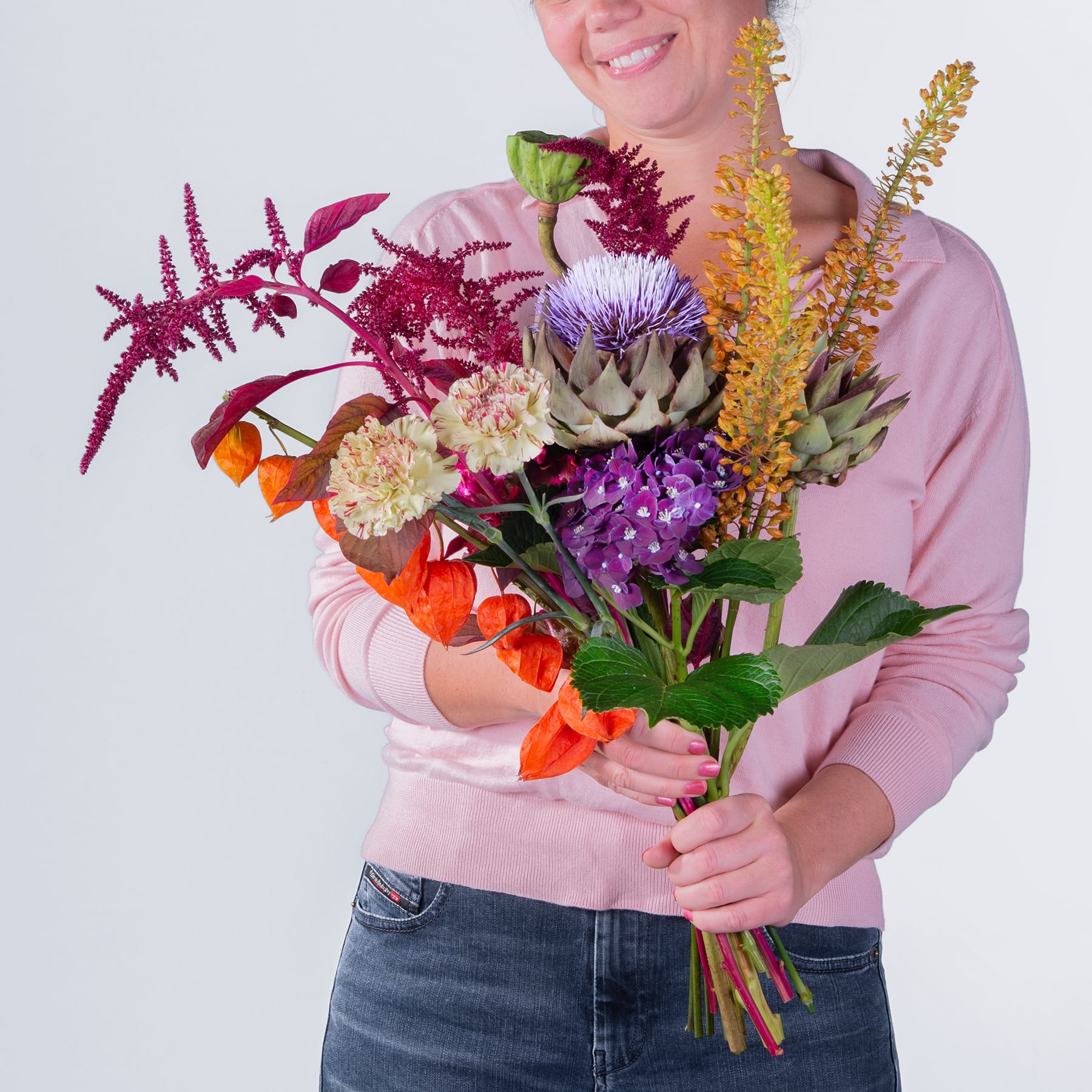 Eine Person in einem rosa Pullover hält vor einem schlichten weißen Hintergrund einen leuchtenden Strauß bunter Blumen, darunter Chrysanthemen, Sukkulenten und andere Herbstblüten.