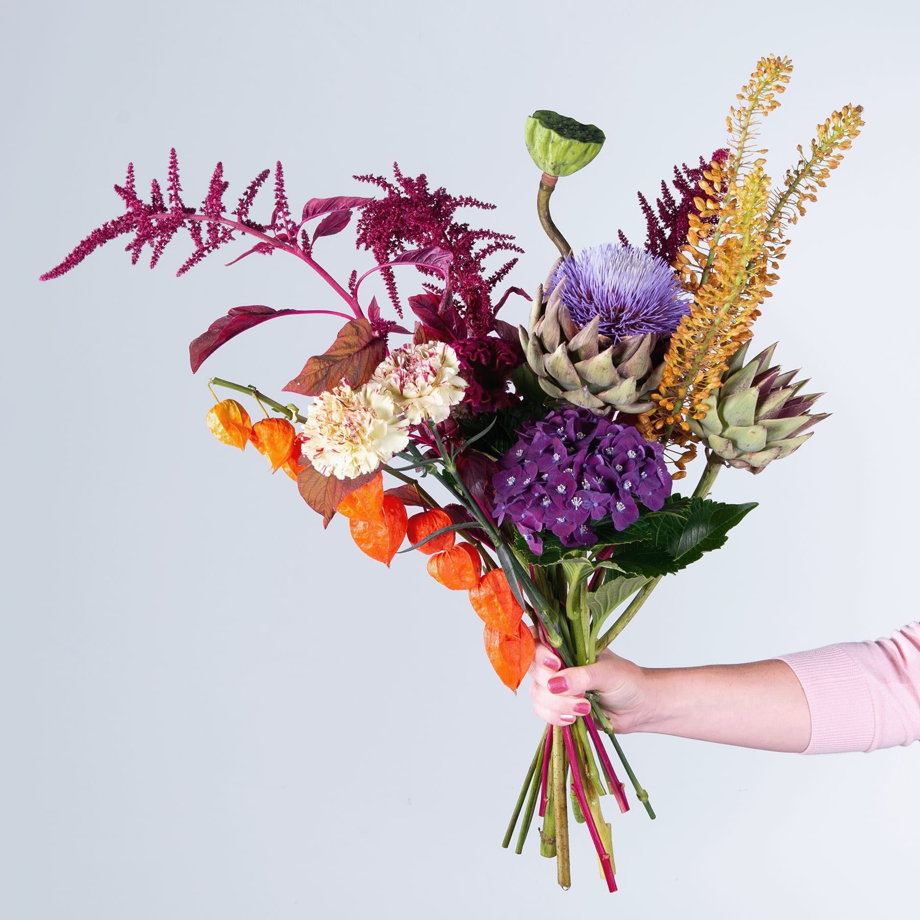 Ein lebendiger Strauß bunter Blumen, darunter violette Artischocken, orangefarbene Ringelblumen und roter Amarant, in einer Hand vor einem schlichten Hintergrund gehalten.