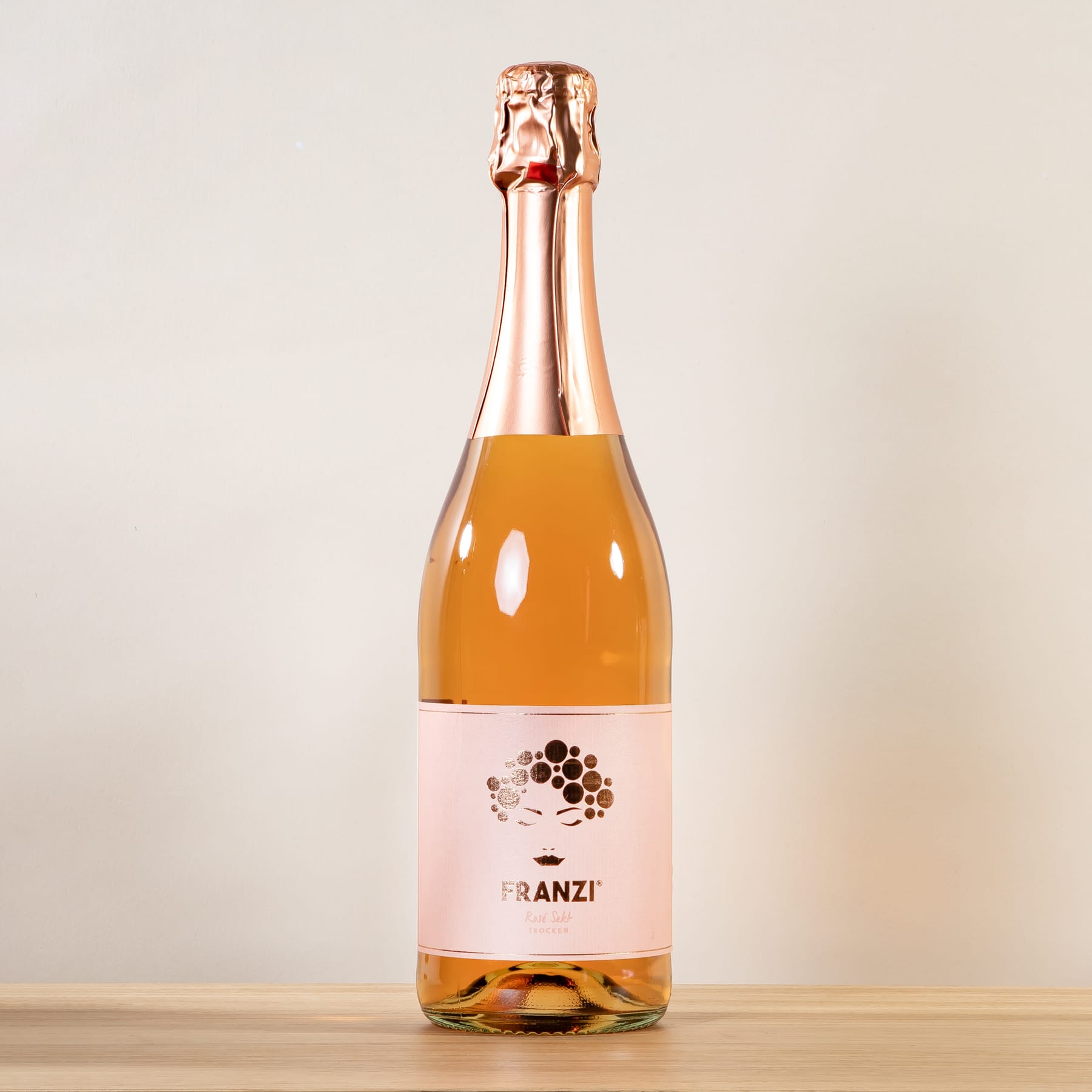 Eine Flasche Franzi-Sekt in Roségold mit Etikett im Wabenmuster auf schlichtem beigem Hintergrund.