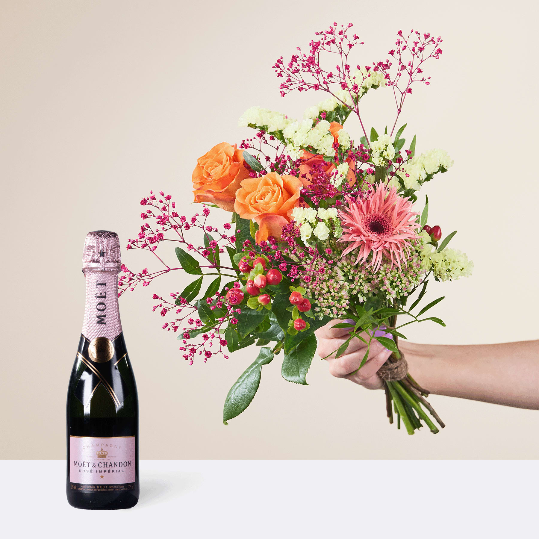 Ein leuchtender Strauß bunter Blumen, darunter orangefarbene Rosen, rosa Gerbera und Grünpflanzen, wird neben einer Flasche Champagner vor einem neutralen Hintergrund in der Hand gehalten.