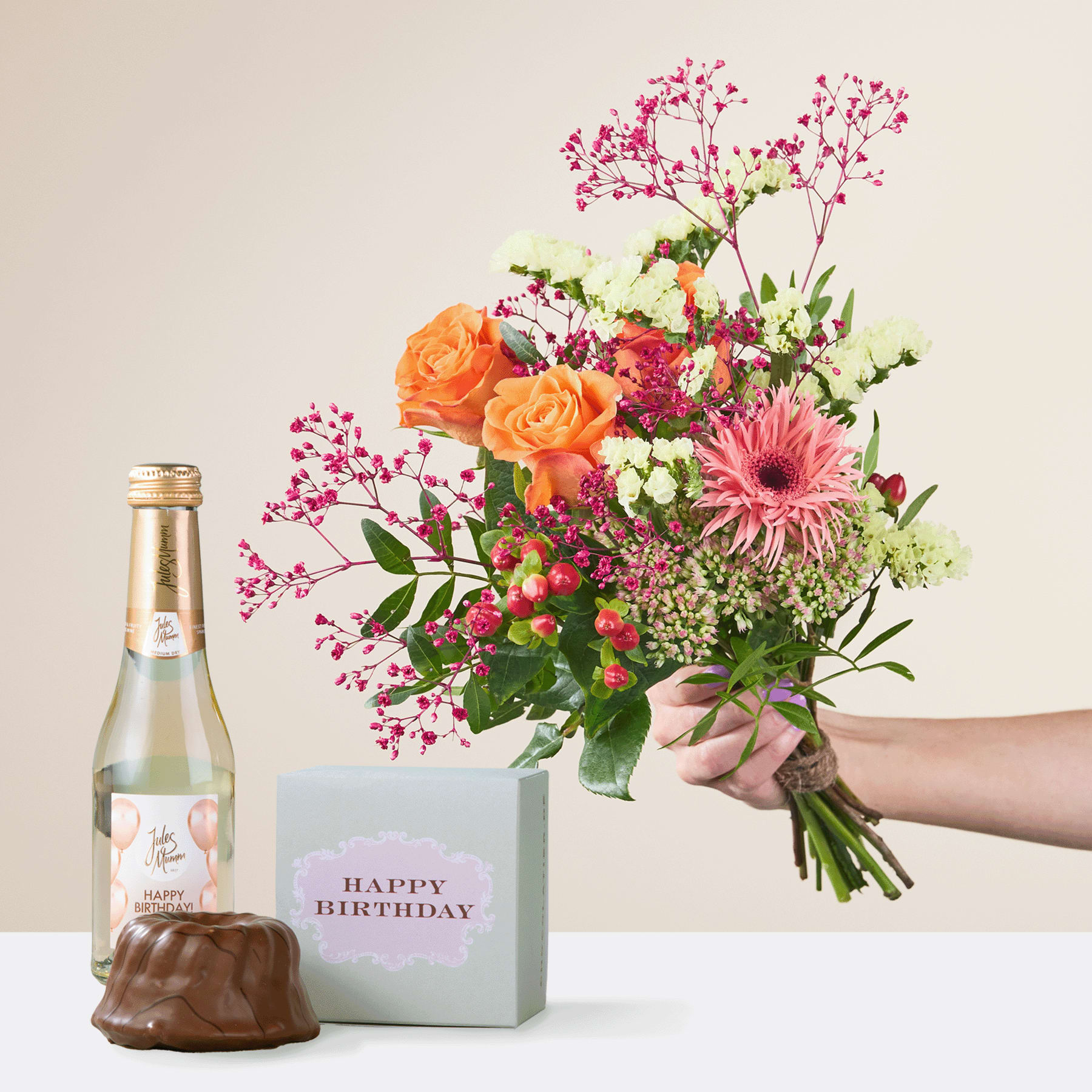 Ein leuchtender Strauß bunter Blumen, darunter orangefarbene Rosen, rosa Gerbera und zartes Schleierkraut, wird zusammen mit einer Geburtstagsgeschenkbox und einer Flasche Champagner in den Händen einer Person gehalten.
