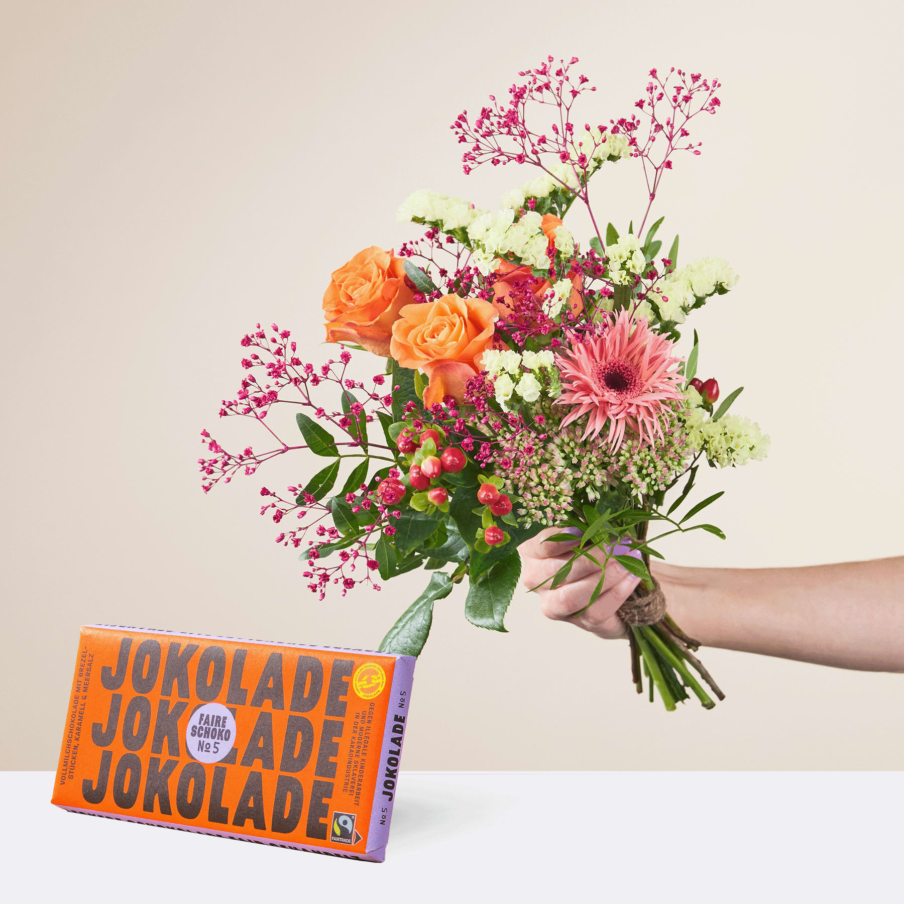 Eine Person hält einen leuchtend bunten Blumenstrauß, darunter orangefarbene Rosen, rosa Gerbera und Grünpflanzen, in der Hand. Im Vordergrund steht auf einer hellen Oberfläche eine Schachtel Jokolade-Schokoriegel.