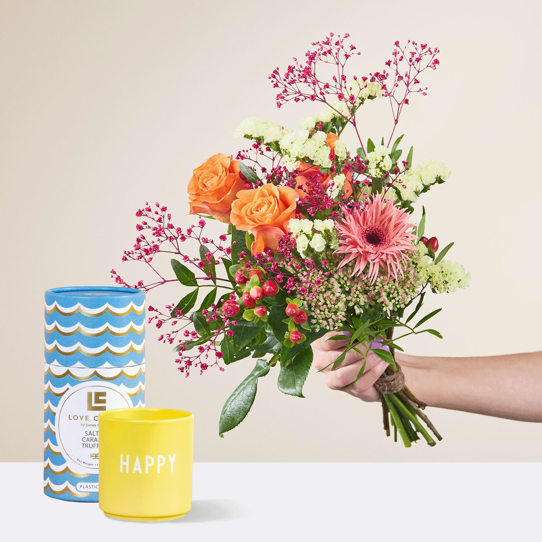Eine Person hält einen leuchtend bunten Blumenstrauß, darunter orangefarbene Rosen, rosa Gerbera und Grünpflanzen, in der Hand. Dazu kommen eine blau-weiß gemusterte Vase und eine gelbe Kerze mit der Aufschrift „Happy“ – alles vor einem schlichten Hintergrund.