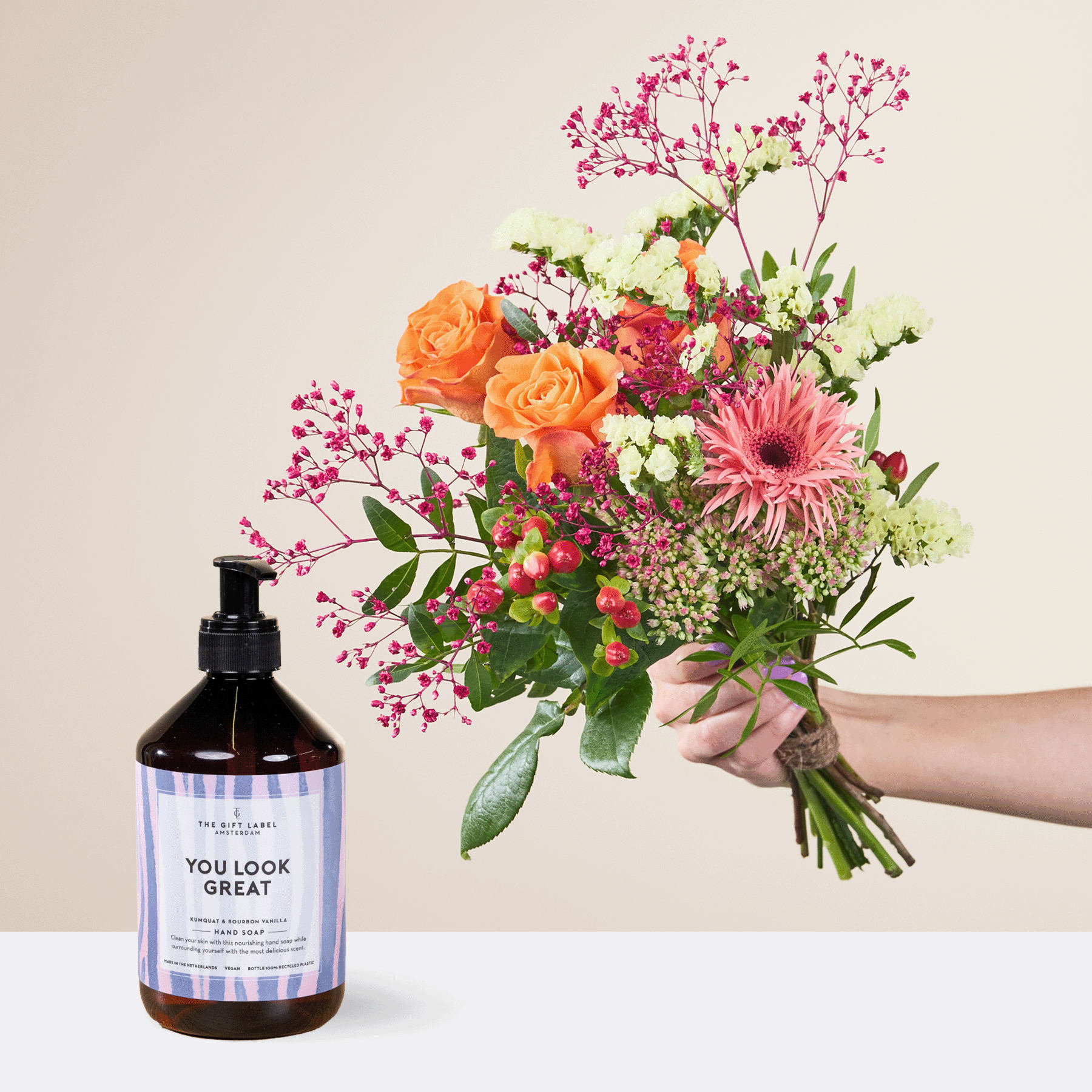 Eine Person hält einen leuchtend bunten Blumenstrauß, darunter orangefarbene Rosen, rosa Gerbera und Grünpflanzen, in der Hand, zusammen mit einer Flasche Handdesinfektionsmittel mit der Aufschrift „Du siehst toll aus“ auf dem Etikett.
