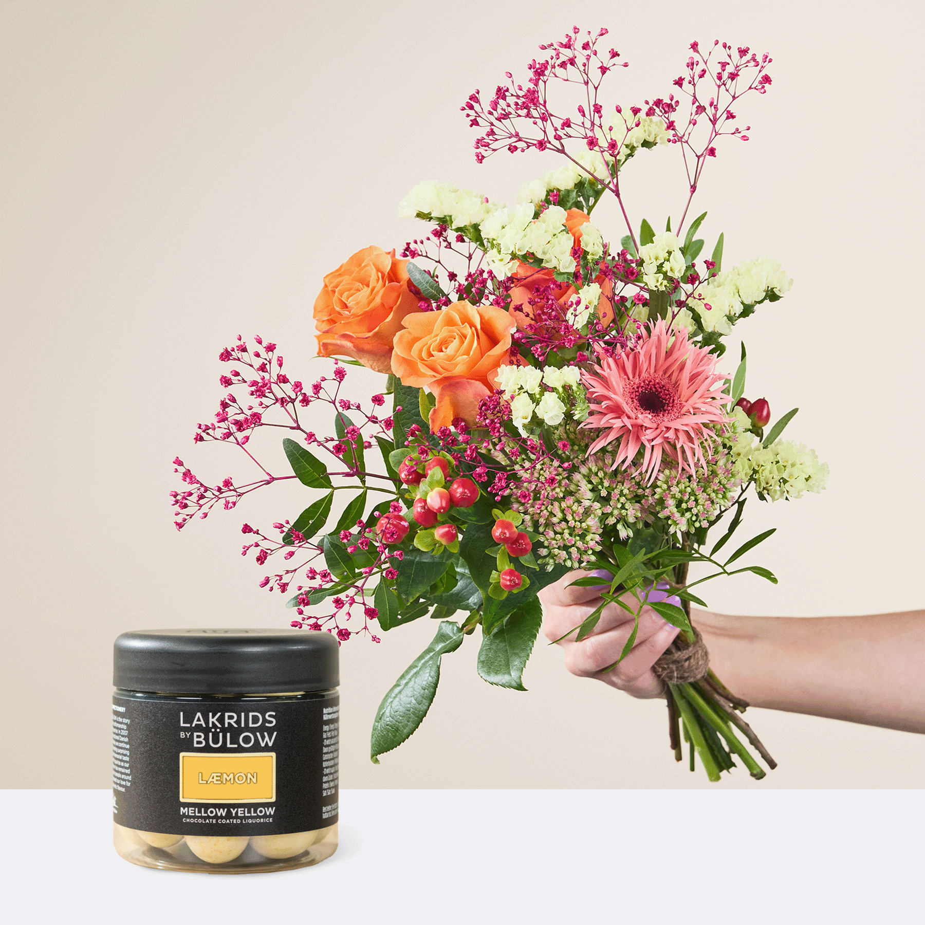 Eine Person hält einen leuchtenden Strauß bunter Blumen, darunter orangefarbene Rosen, rosa Gerbera und zartes Grün, neben einem Glas Lakrids von Johan Bülow in den Händen.