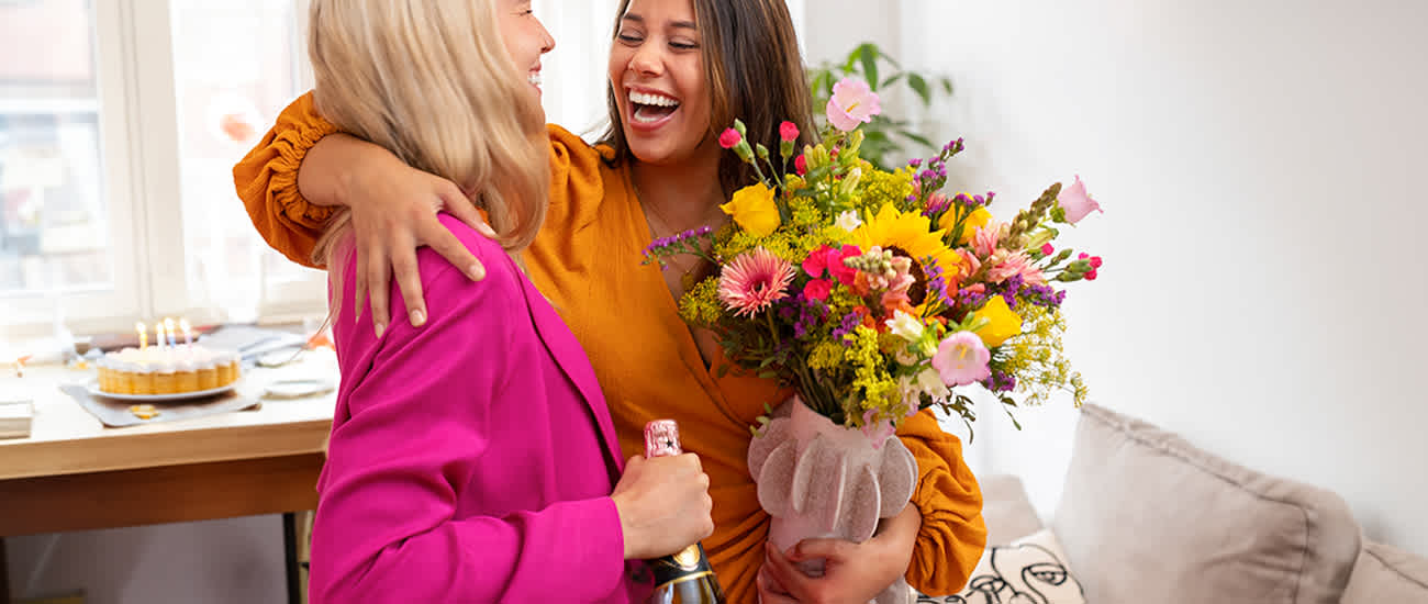 Zwei junge Frauen umarmen sich und lächeln freudig, umgeben von einem leuchtend bunten Blumenstrauß auf einem Tisch im Hintergrund.