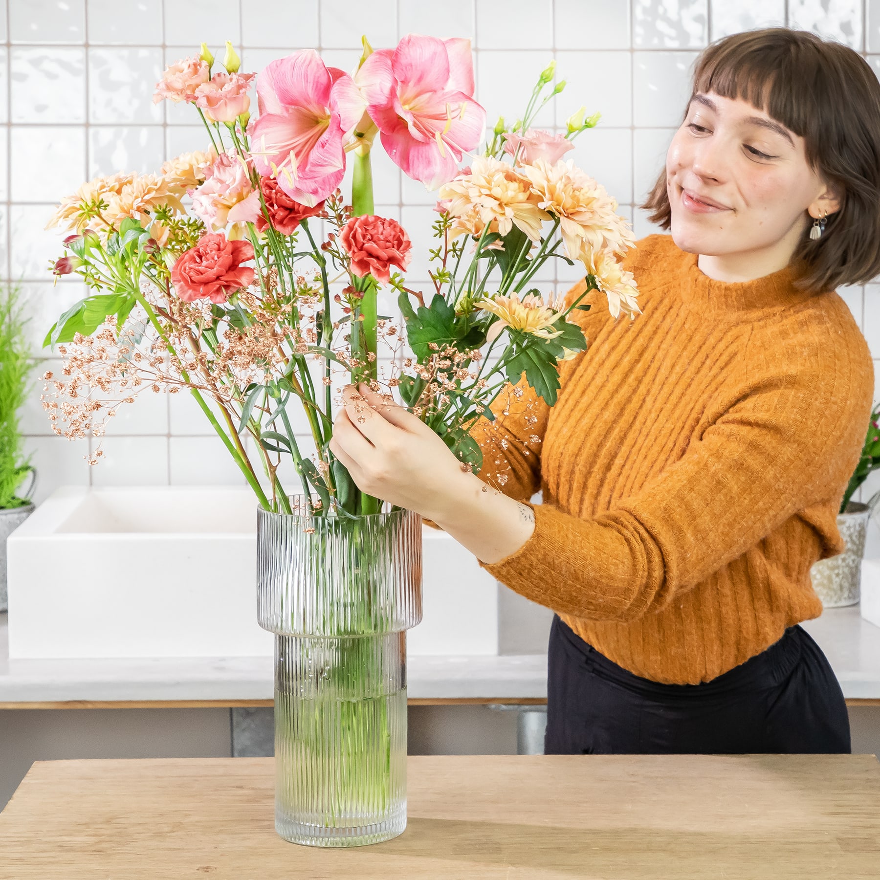 Eine junge Frau in einem senffarbenen Pullover arrangiert einen farbenfrohen Blumenstrauß, darunter rosa und rote Blüten, in einer Glasvase auf einem Holztisch vor dem Hintergrund einer gefliesten Wand.