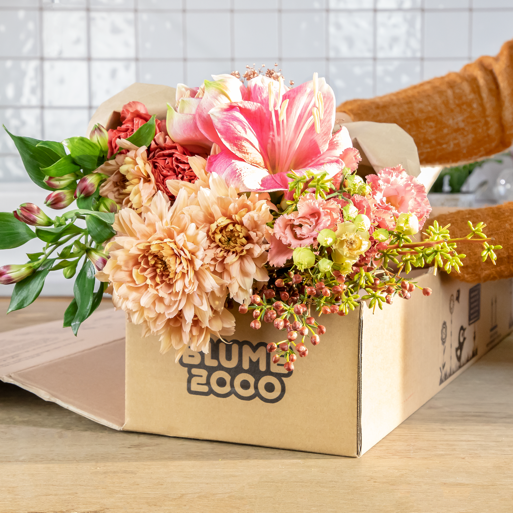Ein leuchtender Strauß bunter Blumen, darunter rosa und gelbe Blüten, steht in einem Karton mit der Aufschrift „Bloom & Eco“ auf einer Holzoberfläche vor einem gefliesten Hintergrund.