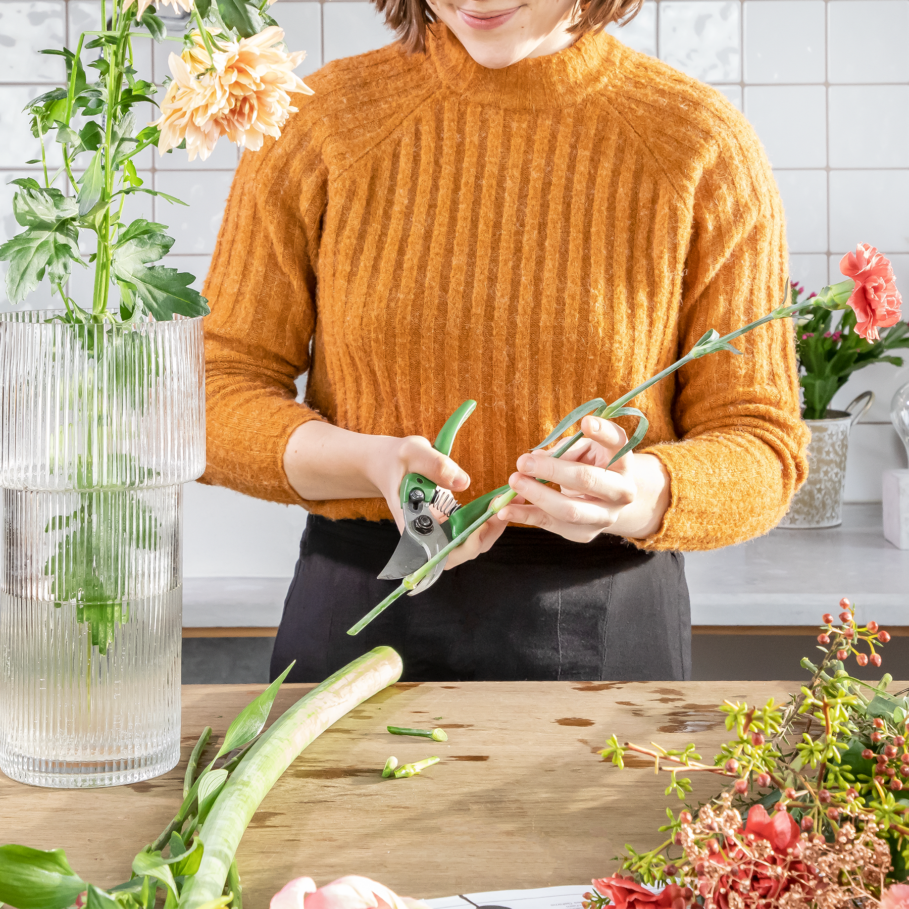 Eine Person in einem warmen, senffarbenen Pullover schneidet in einer Küche, umgeben von Topfpflanzen und Blumen, vorsichtig grüne Stängel.