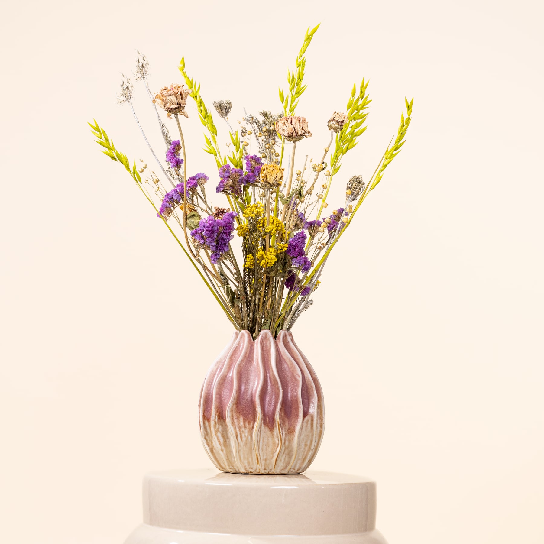 Ein Strauß leuchtend violetter und gelber Wildblumen in einer rosa-weißen Keramikvase steht auf einer schlichten weißen Fläche vor einem hellen Hintergrund.