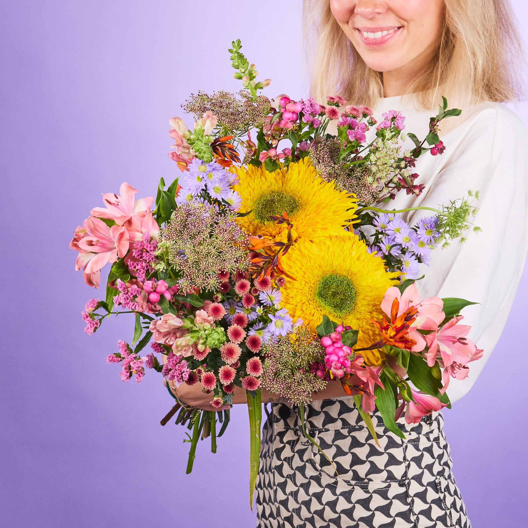 Ein leuchtender Strauß bunter Blumen, darunter gelbe Sonnenblumen, rosa Nelken und verschiedene andere Blüten, wird von einer lächelnden Frau vor einem violetten Hintergrund gehalten.