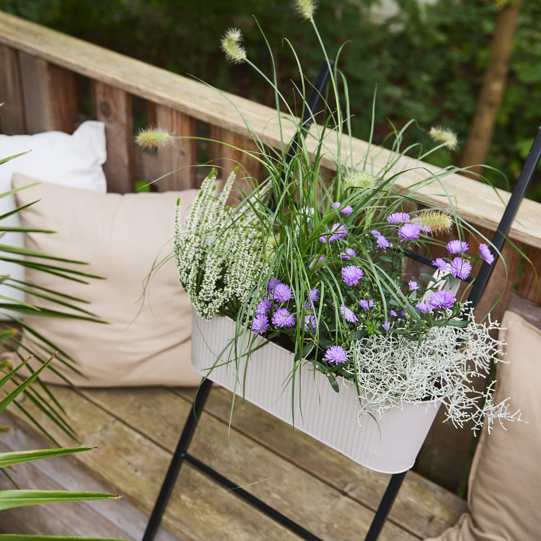 Eine Holzterrasse mit einem Pflanzkasten voller leuchtender lila und weißer Blumen, umgeben von üppigem Grün und einem gemütlichen Sitzbereich im Freien.