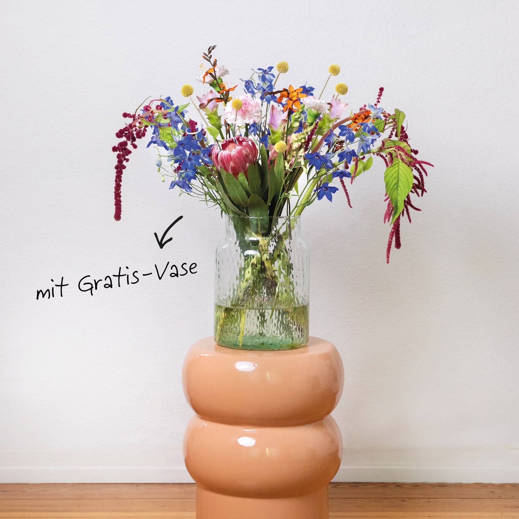 Ein leuchtender Strauß bunter Blumen, darunter Tulpen und andere Blüten, wird in einer Glasvase präsentiert, die auf einer Holzoberfläche vor einem schlichten weißen Hintergrund steht.