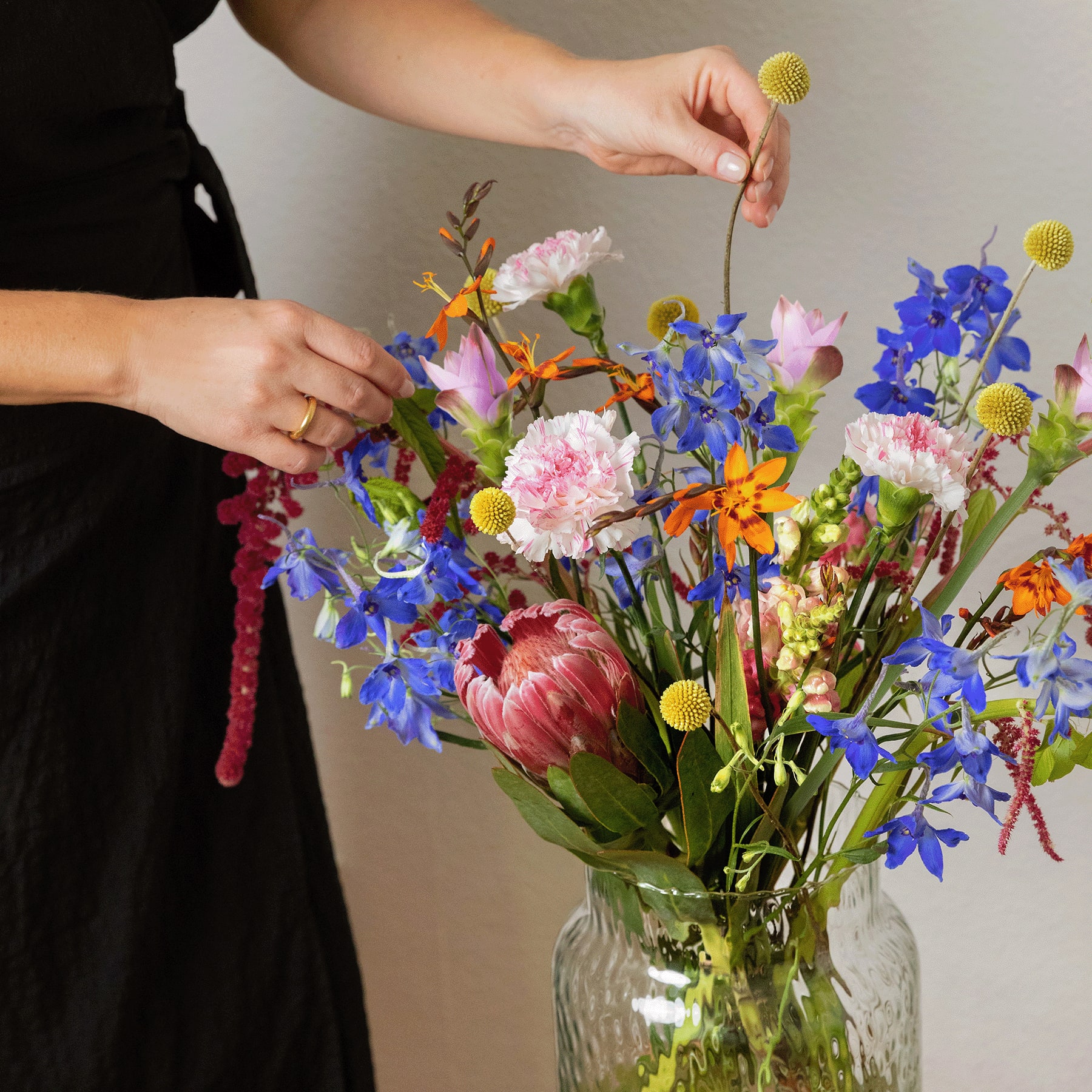 Ein lebendiger Strauß bunter Blumen, darunter Pfingstrosen, Gänseblümchen und Schwertlilien, steht in einer Glasvase vor einem schlichten Hintergrund.