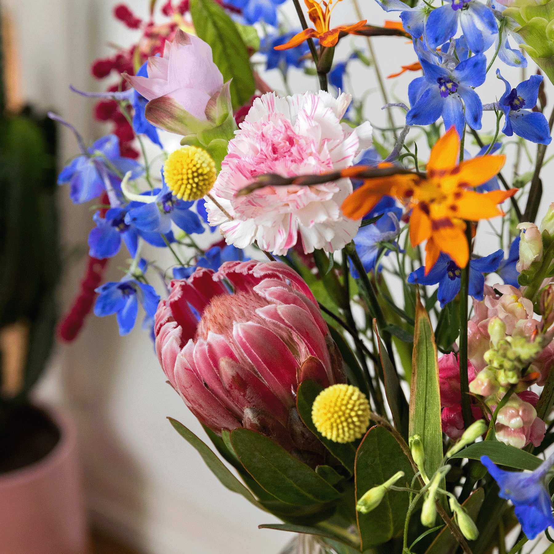 Ein lebendiger Strauß bunter Blumen, darunter rosa Pfingstrosen, blaue Rittersporne und gelbe Knopfblumen, arrangiert in einer Vase vor einem unscharfen Hintergrund.